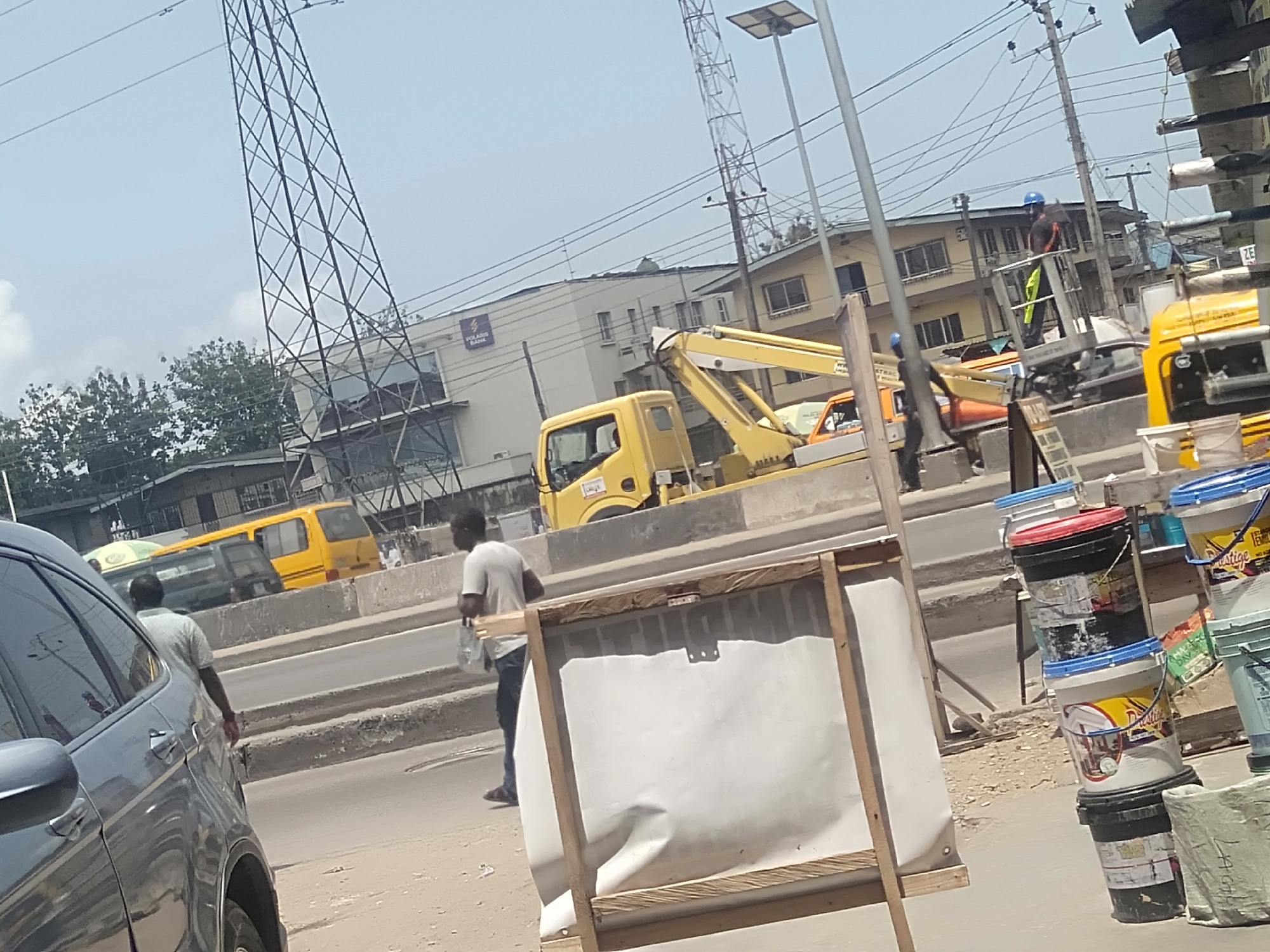 The image shows a street scene in Shangisha, Nigeria. A gray car is in the foreground, partially obstructing the view. Two men are walking on the street, one closer to the car wearing a gray shirt and carrying a bag, and the other further away, also wearing a gray shirt.
In the background, a yellow bucket truck with its arm extended is present with a worker in the bucket. There is a yellow bus on the road and a Polaris Bank building can be seen in the distance. Construction work is being done on top of a building behind Polaris Bank.
Stacked paint buckets are visible on the right side of the image, and a white advertisement board on a wooden frame is in the foreground. Utility poles and power lines are seen throughout the scene, and a tall lattice transmission tower is visible behind the buildings.