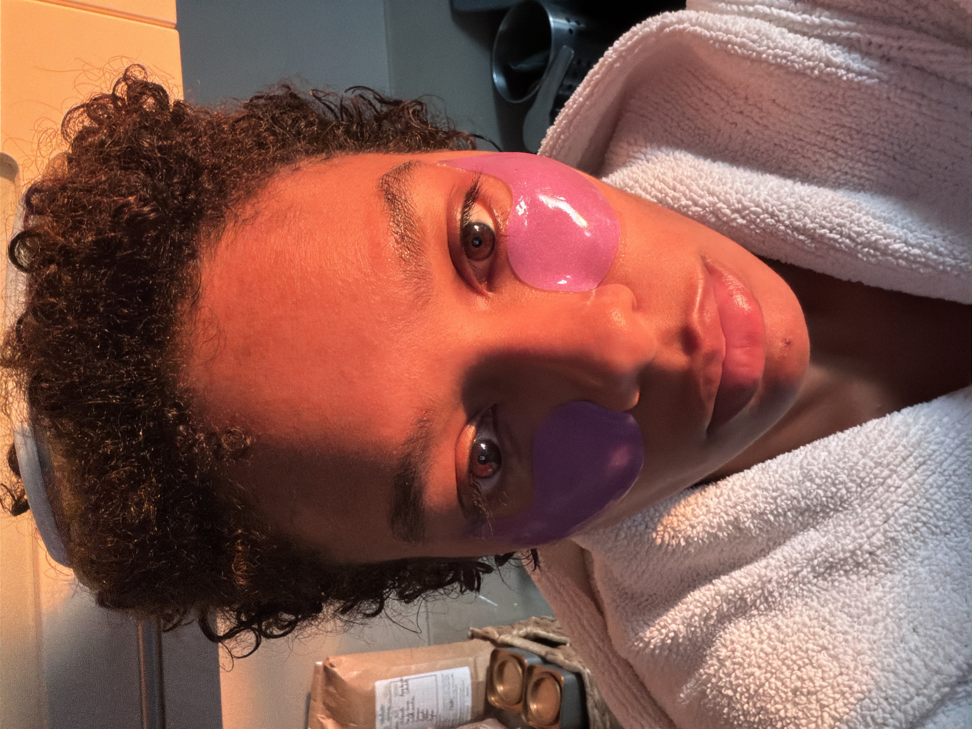 The image shows a close-up, head-on shot of a woman's face, illuminated by warm, orange-toned light, likely from a sunrise or sunset, which casts dramatic shadows and highlights across her features. The woman has dark, curly hair pulled up, and is wearing purple under-eye masks. She is also wearing a white, terrycloth robe. In the background, to the left, is a brown paper bag and some small metal containers, possibly part of a beauty or skincare routine. To the right, a dark object, possibly a mug or cup, is visible. The overall impression is one of a personal moment of self-care, perhaps in a domestic setting like a bathroom or kitchen, with the strong backlight creating an intimate and stylized atmosphere. The specific location in Champs-sur-Marne, France, is not discernible from the visual elements of the image itself.