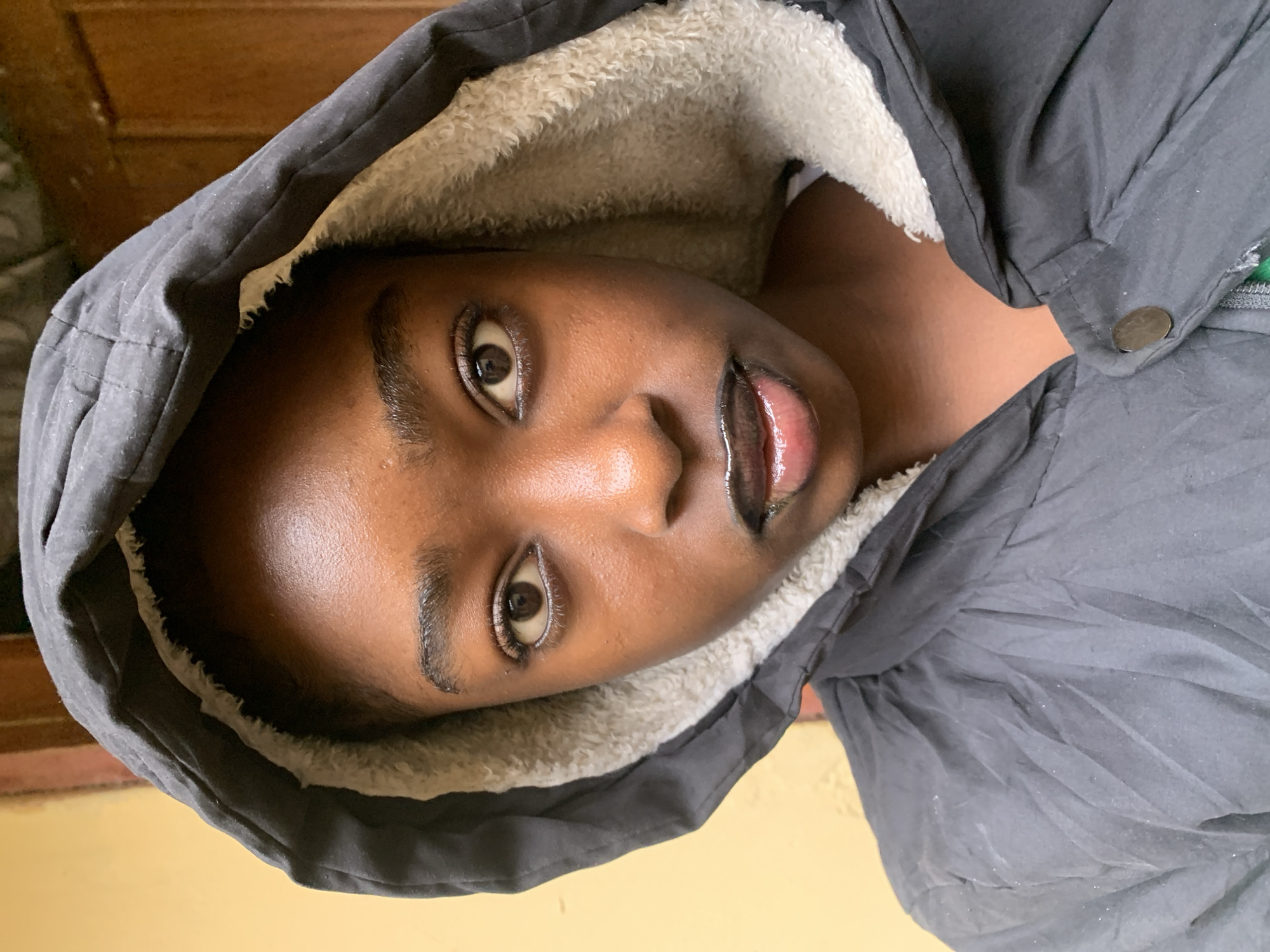 A close-up, head-and-shoulders portrait of a young Black person is shown. They are wearing a dark gray hooded jacket with a light-colored, fuzzy lining. The hood is pulled up, framing their face. Their skin is dark and appears slightly shiny. They have dark, wide-set eyes, dark eyebrows, and dark hair. Their lips are full and painted with a dark, glossy lipstick. The background is indistinct, with a vertical element that appears to be a wooden door on the right and a light-colored wall on the left. The image appears to have been taken indoors or in a sheltered outdoor space. No specific location in Rafin Par, Nigeria is discernible from the image.