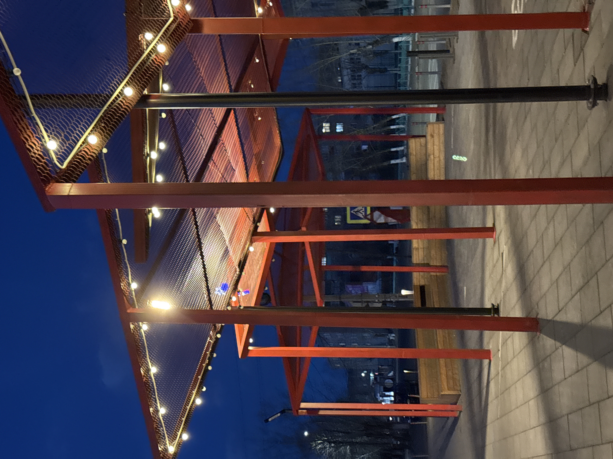A low-angle, rotated perspective captures an open-air structure, possibly a covered pedestrian area, illuminated by string lights strung across its metal framework. The framework consists of red metal beams supporting a mesh roof. Beyond the structure, one can discern buildings and street elements indicative of an urban environment. The sky is deep blue, suggesting twilight or night. The pavement below is light gray and made of brick. Buildings in the background contain visible windows. A street sign is seen beneath the beams. The location is likely in Borzya, Russia, although there is no specific indicator present in the image that confirms this.