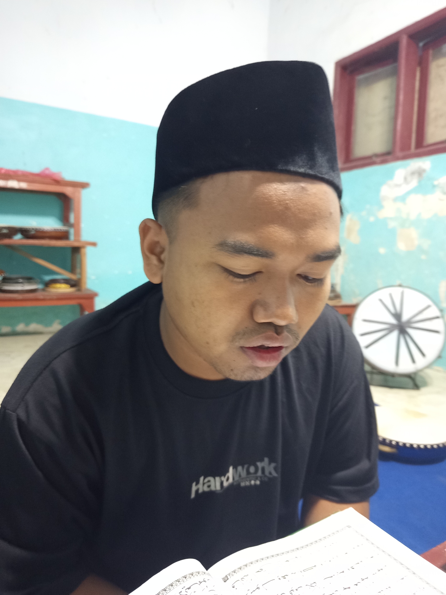 A man wearing a black peci hat and a black t-shirt with "HardWork" printed on it is reading a book in a room. The room has light blue walls and a wooden shelf with items on it. A round object with spokes is visible in the background. The man is looking down at the book. The scene appears to be in a residential setting in Ngablak, Indonesia.