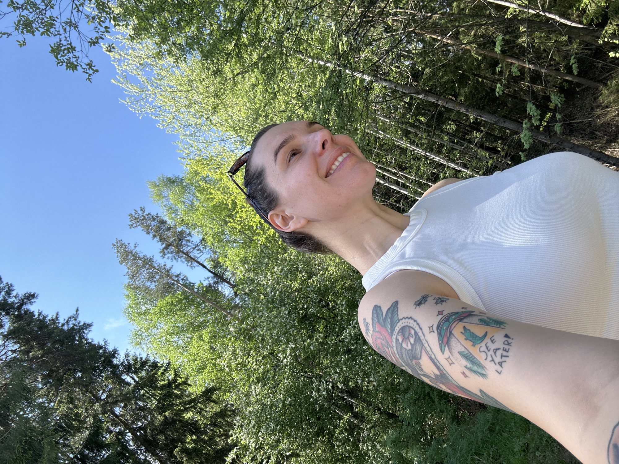 The image is a low-angle selfie of a woman with fair skin, taken outdoors, likely in a forested area. The woman is smiling and looking upward and to the right. She is wearing a white tank top and has sunglasses resting on her head. She has a tattoo on her upper left arm. The background consists of green trees and a blue sky. The location may be Прикамский, Russia.