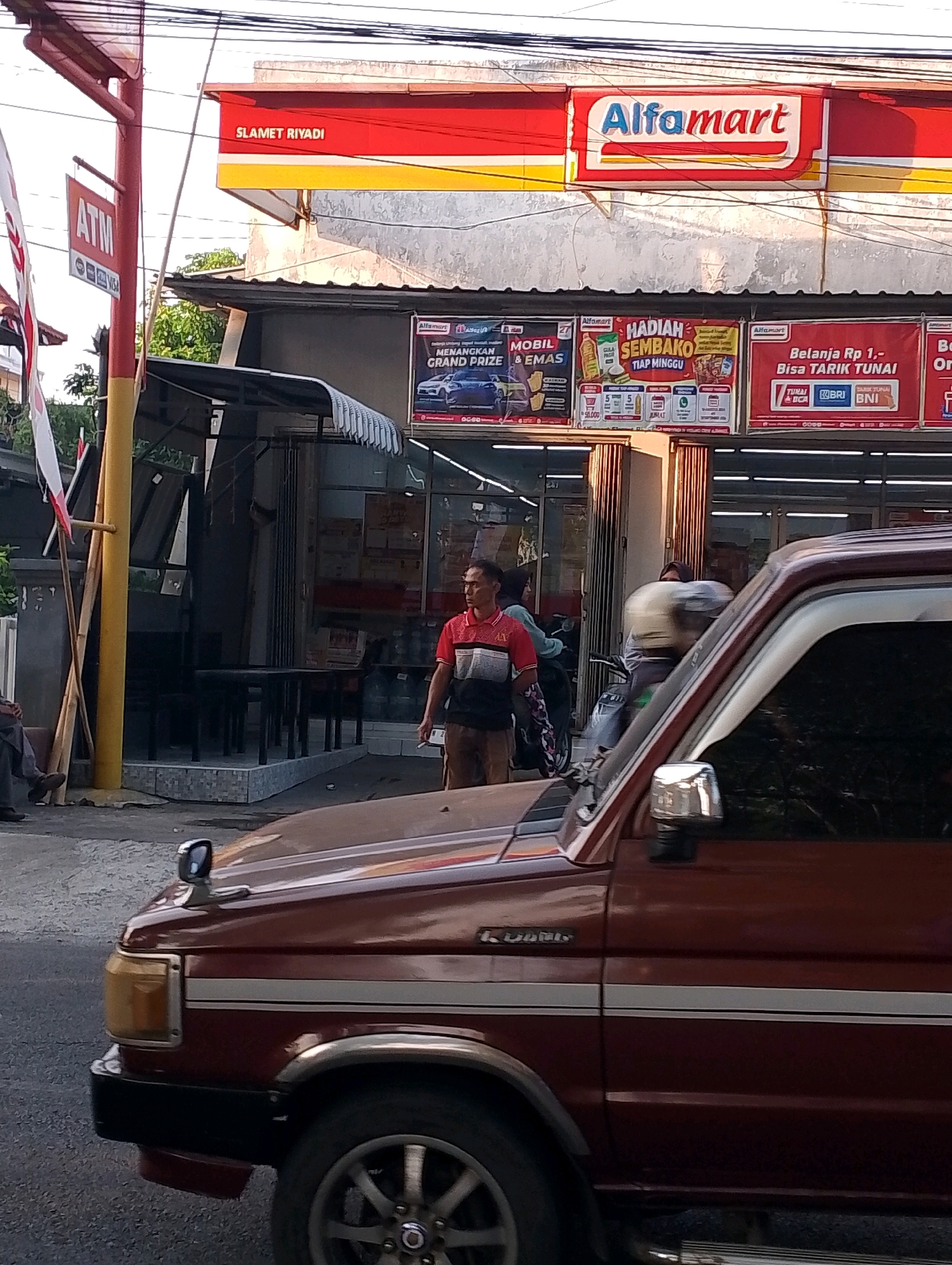 The image shows a street scene in Probolinggo, Indonesia, with a prominent Alfamart convenience store. A dark red SUV is parked in the foreground, partially obscuring the view of the store and its surroundings. The Alfamart store has a red and yellow awning with the store's logo clearly visible. Beneath the awning, several advertisements are displayed, promoting various offers and products, including a "Hadiah Sembako" (grocery gift) promotion and a "Belanja Rp 1,- Bisa Tarik Tunai" (Shop for Rp 1,- Can Cash Out) offer.
To the left of the Alfamart, a red pole with an "ATM" sign and logos for Visa is visible, suggesting an ATM is available. A man in a red and black striped shirt and brown pants is standing in front of the store, holding a cigarette. Other individuals and a motorcycle are also present near the store entrance, indicating typical street activity. In the background, there are trees and buildings, characteristic of an urban environment in Indonesia. The overall lighting suggests it is daytime, possibly late afternoon, with sunlight casting shadows.