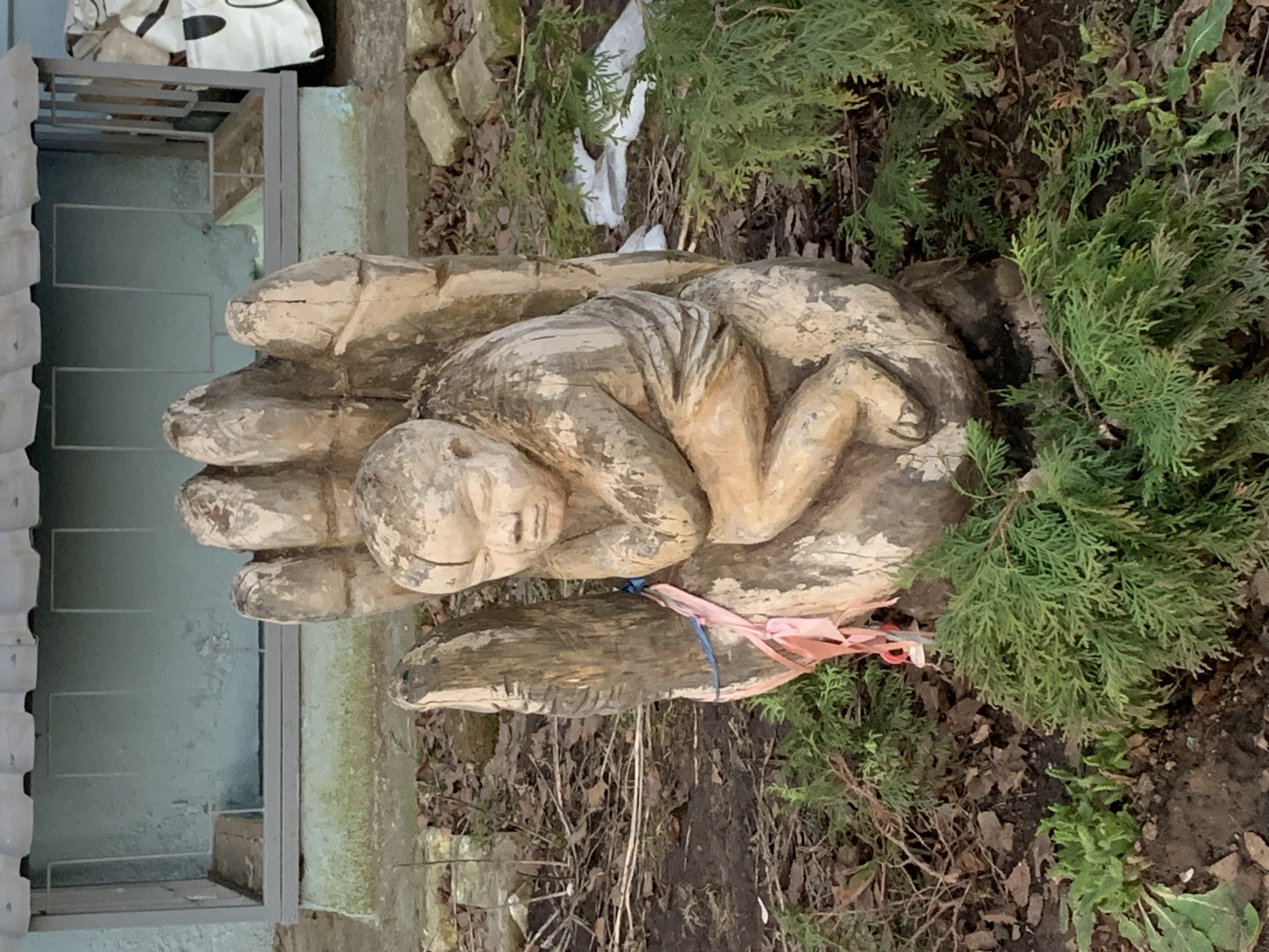 The image depicts a carved wooden sculpture of a hand cradling a fetus. The sculpture is located outdoors near a building, possibly in Chaikovsky, Russia. The sculpture is weathered and partially surrounded by evergreens, dry leaves, and other plants. A pink ribbon is tied to the base of the sculpture near the wrist of the hand. The sculpture is positioned on what appears to be dirt and dead leaves.