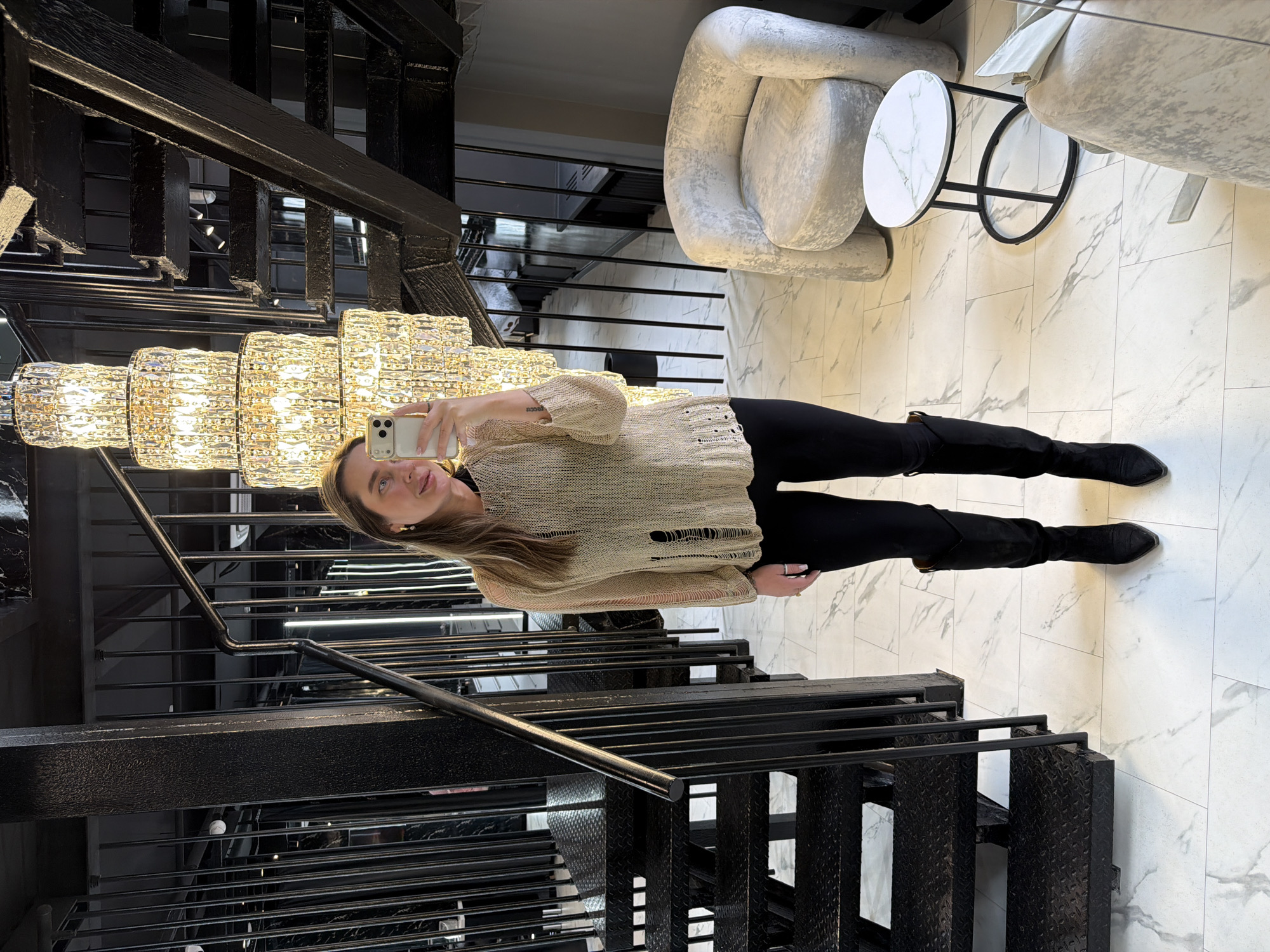 A full-length shot captures a woman taking a selfie in what appears to be an interior space, possibly a commercial or residential building. The setting is Moscow, Russia. The woman, positioned horizontally in the frame, holds a smartphone to take the picture. She is dressed in black leggings and boots, paired with a light-colored, distressed sweater.
She stands near a prominent black metal staircase, which adds an architectural element to the scene. Above her, a modern, multi-tiered chandelier illuminates the area. To the right, marble-patterned flooring and partial views of modern furniture, including an arm chair and a small round table, are visible. The scene implies a contemporary, well-designed interior.