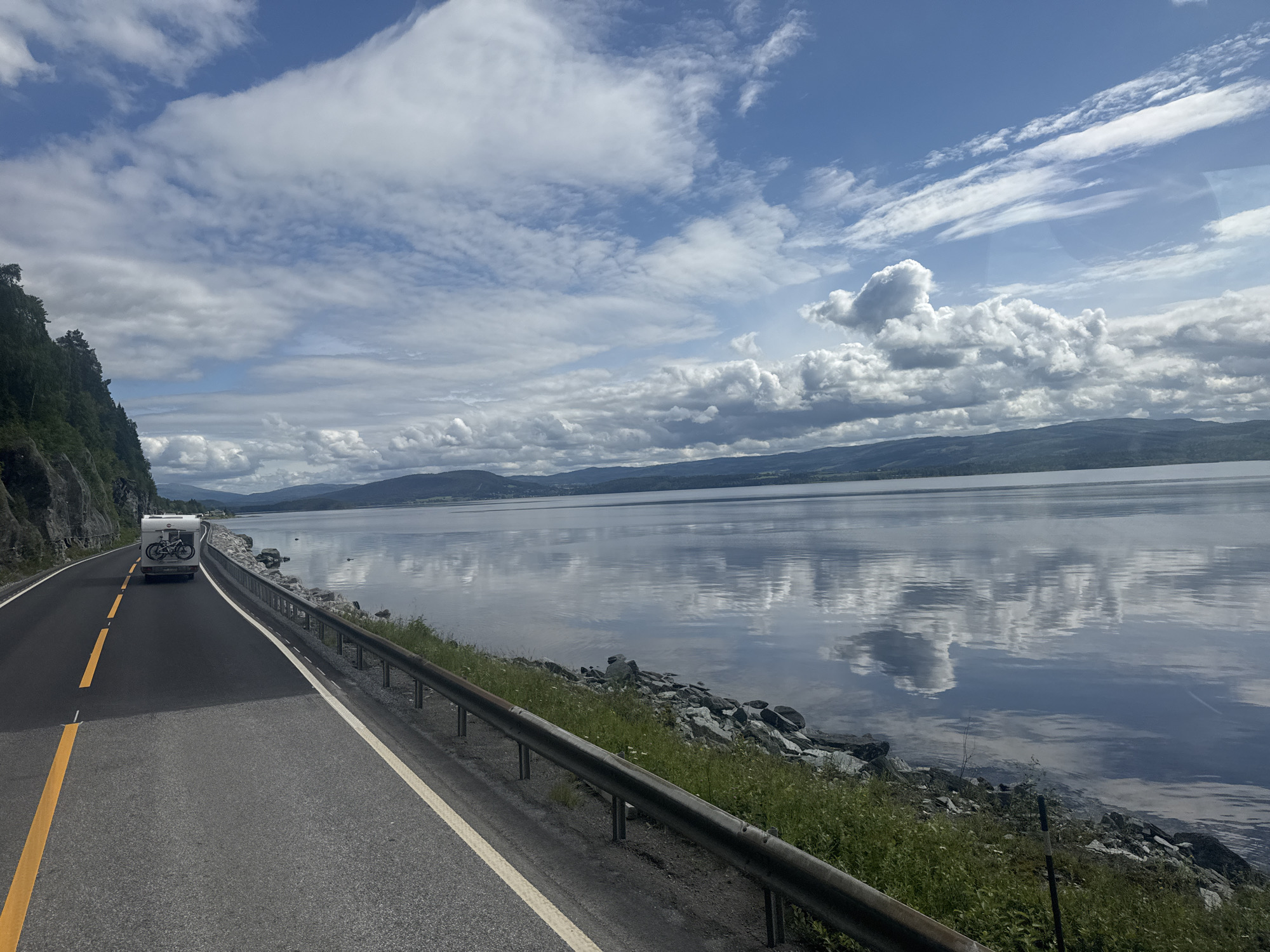 A motorhome with bicycles mounted on the rear travels along a paved road that curves to the left. The road is bordered by a sheer rock face on the left and a metal guardrail and rocky shoreline on the right. The shoreline is covered in grass and scattered rocks. Beyond the shoreline is a large, calm body of water that reflects the sky and clouds. The sky is partly cloudy with a mix of blue sky and large, white cumulus clouds. In the distance, rolling hills form the horizon. The scene appears to be located in Snåsa, Norway, given the natural landscape and the presence of a fjord or large lake.