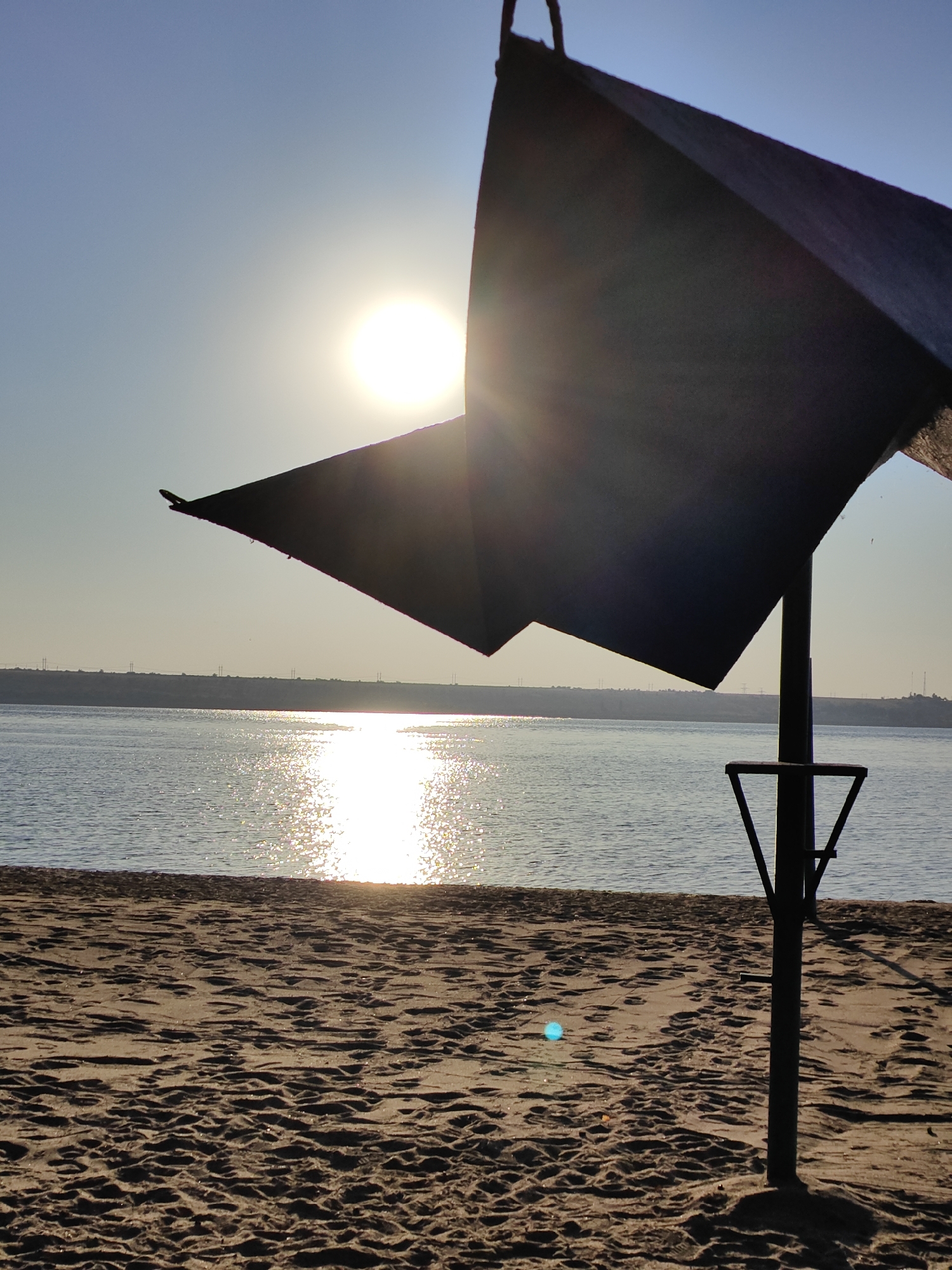 A sun umbrella and its pole are silhouetted against a bright sun setting over a body of water. The sand of a beach is in the foreground, with the water and a distant, hazy shoreline in the background. The umbrella is dark and angular, with a triangular shape visible. The sun is high in the sky, its rays glinting off the water's surface. There are no people or animals visible in the image. The location is identified as Nezavertaylovka, Moldova.