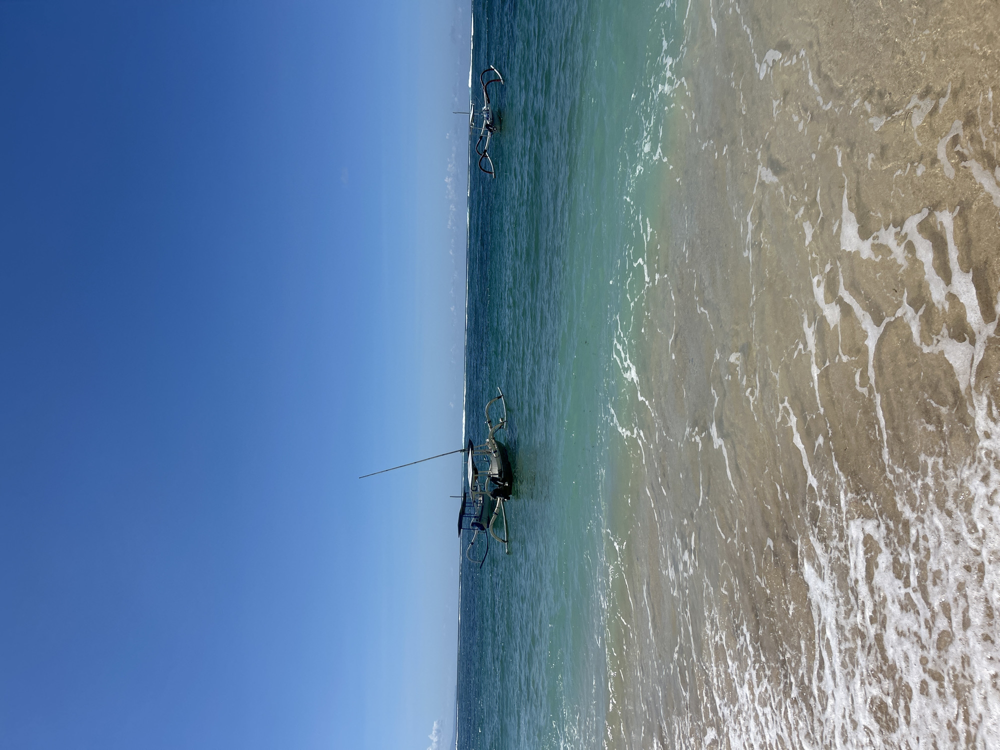 The image depicts a beach scene in Kuta Selatan, Indonesia. Two traditional Indonesian fishing boats, known as Jukung, are visible in the water. The boats have outriggers and a slender mast. The water is a mix of turquoise and light brown near the shore where the waves are breaking. The sky is a clear, bright blue.