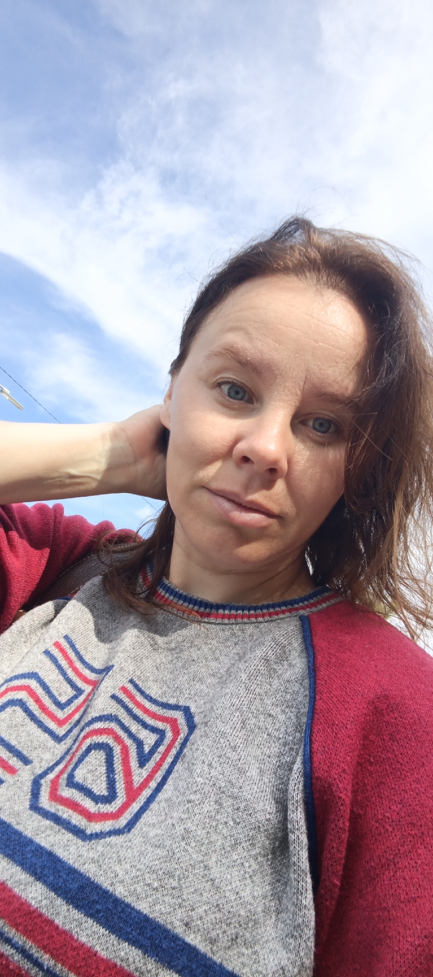 A medium shot taken from a low angle shows a woman with light brown hair and blue eyes looking directly at the camera. She is outside under a blue sky with some clouds. Her right hand is behind her head, and she is wearing a sweater. The sweater is gray on the left side and red on the right side, with a blue and red striped collar. The gray portion of the sweater has a design with red and blue lines forming a shield-like shape. The background is dominated by the sky, with a hint of some infrastructure on the far left. The image was taken in Chaikovsky, Russia.