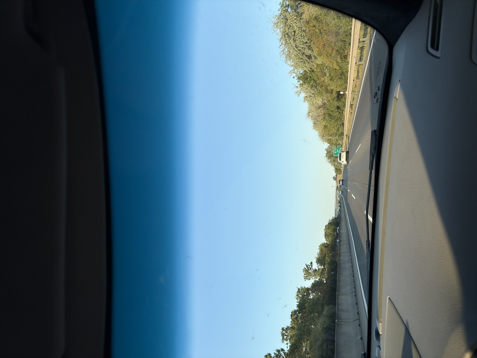 This media file depicts a view from inside a vehicle traveling on a highway. The perspective is looking forward and slightly to the right. The sky occupies a significant portion of the frame, transitioning from a deep blue on the left to a lighter blue on the right. The highway is visible in the distance, with multiple lanes and vehicles, including cars and a white van. Trees and foliage line the sides of the highway, appearing green and lush. A concrete barrier separates the highway from the trees on the right side of the frame.
The time of day appears to be daytime, likely late morning or afternoon, given the bright sunlight and clear sky. The weather seems fair and sunny. There are some faint specks on the windshield, possibly dust or water spots.
Notable details include road signs, such as a green highway sign with white lettering that is partially visible in the distance. The interior of the vehicle is visible on the right side of the frame, showing a grey dashboard and part of the windshield. The overall impression is one of a scenic drive on a clear day.