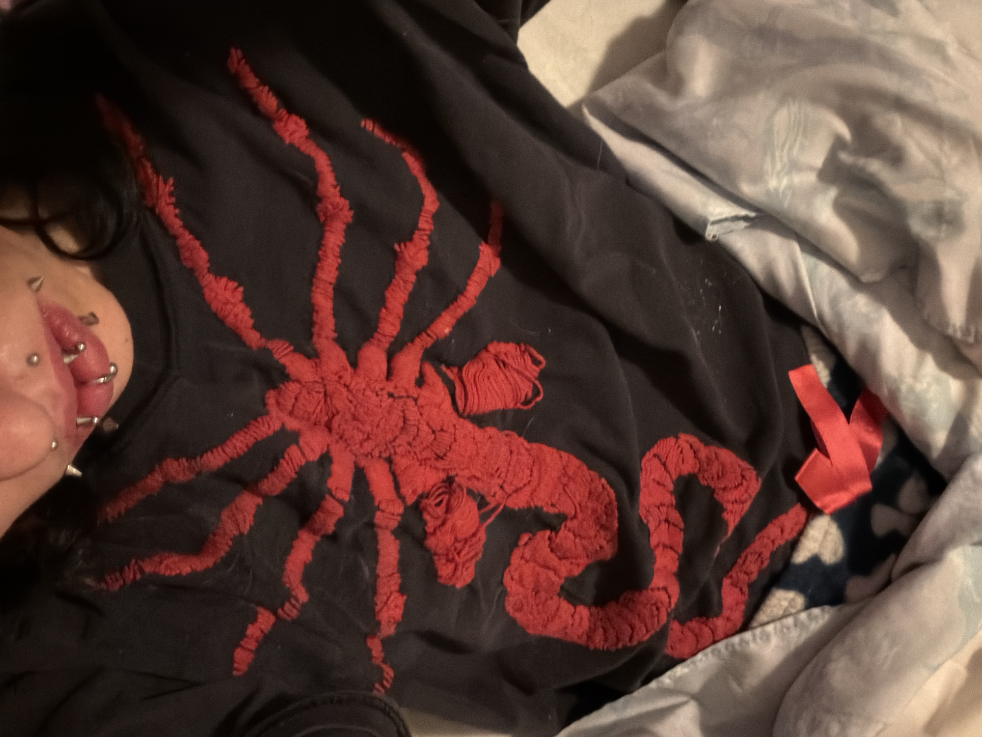 An individual with multiple facial piercings is lying in bed, wearing a black t-shirt with a large, red, textured design of a spider or octopus-like creature. The bed is covered in gray and white patterned sheets. A red ribbon is partially visible near the edge of the shirt. The location in Sofia, Bulgaria, cannot be determined from the image.