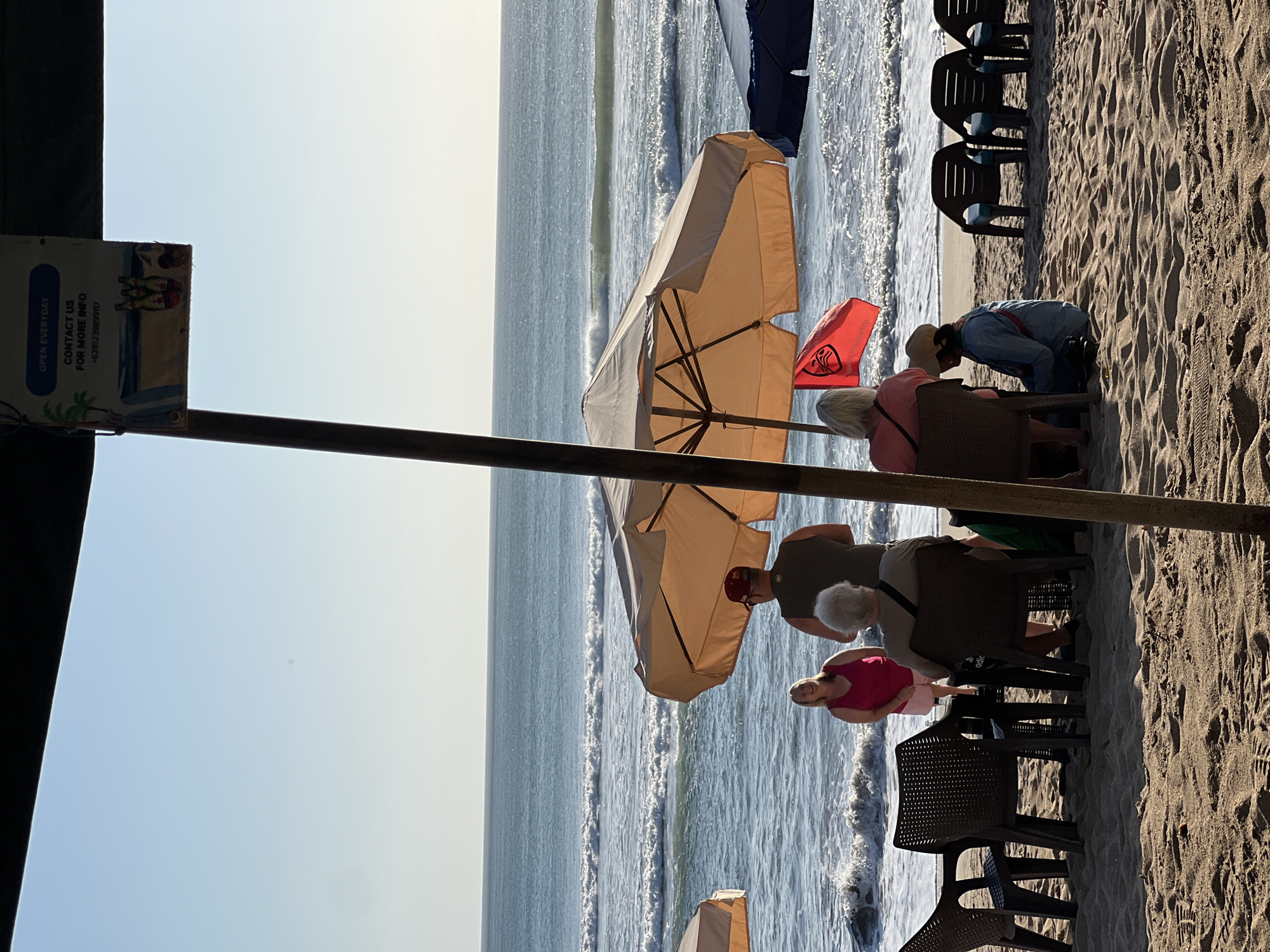 The image depicts a beach scene in Kuta, Indonesia, likely during the day due to the bright sunlight. Several people are visible, some seated at tables under a large tan umbrella, while others stand or sit on the sand. The ocean with waves is in the background, and the sky is a clear, pale blue.
Specifically, there are at least six individuals visible. Two people are seated at a table under the umbrella, facing away from the camera, with one person wearing a light-colored hat. Another person, wearing a pink top and white shorts, stands by the water's edge, smiling. A man wearing a dark t-shirt and a red baseball cap stands next to her, facing the ocean. Further to the left, two dark plastic chairs are empty at a table. On the right side of the frame, three dark plastic lounge chairs are visible, also empty. A red flag with a white logo is attached to a pole under the umbrella. In the foreground, the sandy beach shows footprints. A sign hanging from a pole above the umbrella indicates "OPEN EVERYDAY CONTACT US FOR MORE INFO +6281239899117" and features illustrations of palm trees and bottles. The overall atmosphere suggests a relaxed beachside establishment.