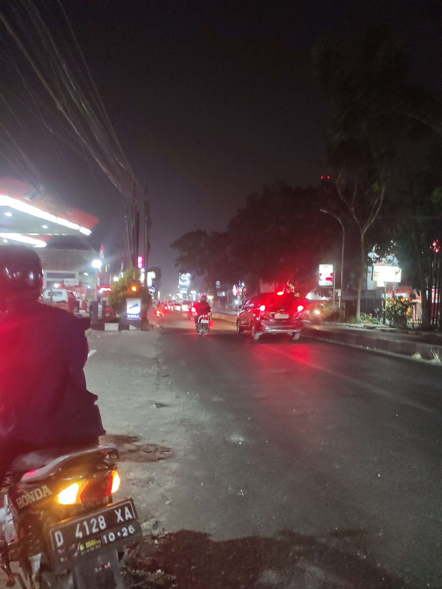 The image captures a night scene on a street in Kertamulya, Indonesia, likely near a gas station based on the prominent canopy structure on the left. A person wearing a dark jacket and helmet is partially visible in the foreground, seated on a motorcycle. The motorcycle's taillight is illuminated, and a license plate reading "D 4128 XA 10.26" is clearly visible. The street is wet, indicating recent rain, and reflects the various lights from passing vehicles and streetlights. Several cars and motorcycles are seen on the road, with their headlights and taillights creating streaks of light in the darkness. Trees and some buildings line the side of the street. The overall atmosphere is that of a typical urban night scene, with traffic and ambient lighting.