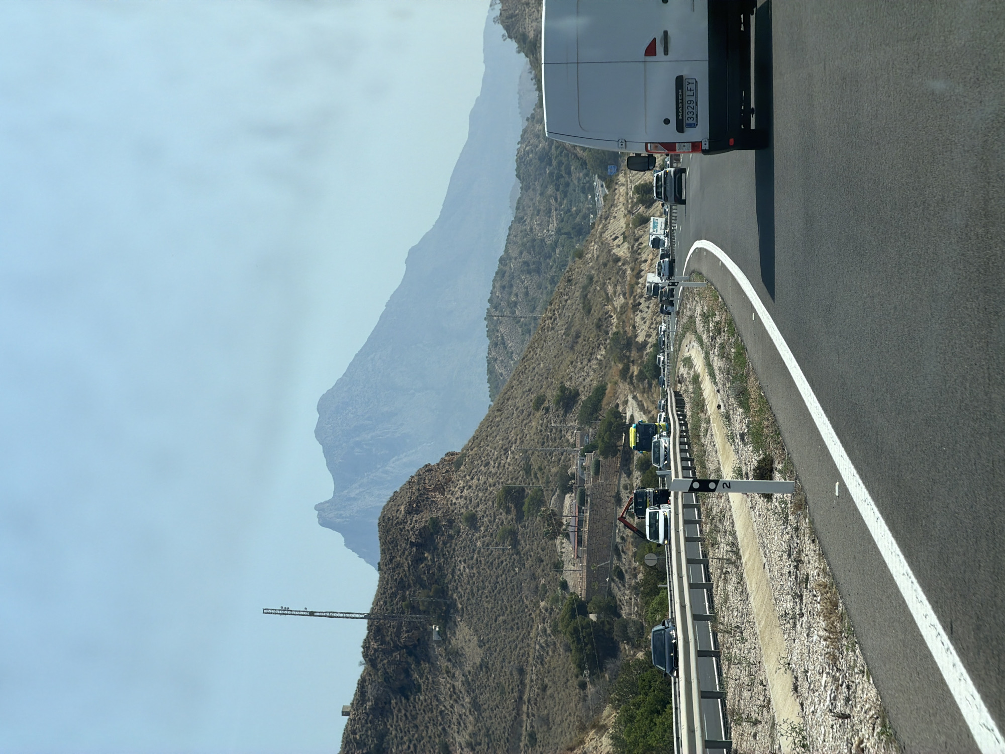 The image depicts a highway scene in Villajoyosa, Spain, under a clear, hazy sky. A white van, possibly a Renault Master based on the visible text on its license plate, is prominently in the foreground, traveling on the right side of the road. The van's license plate reads "3329 LFY". The highway is a multi-lane asphalt road with white lane markings. On the left side of the highway, there is a dry, hilly landscape characterized by sparse vegetation and rocky terrain. A tall communication tower is visible on one of the hills, along with a small structure nearby. In the distance, a large, rugged mountain dominates the horizon, its peak shrouded in a light haze. Several other vehicles, including cars and trucks, are visible on the highway ahead, indicating moderate traffic. The overall impression is a sun-drenched, arid environment typical of the Mediterranean region.