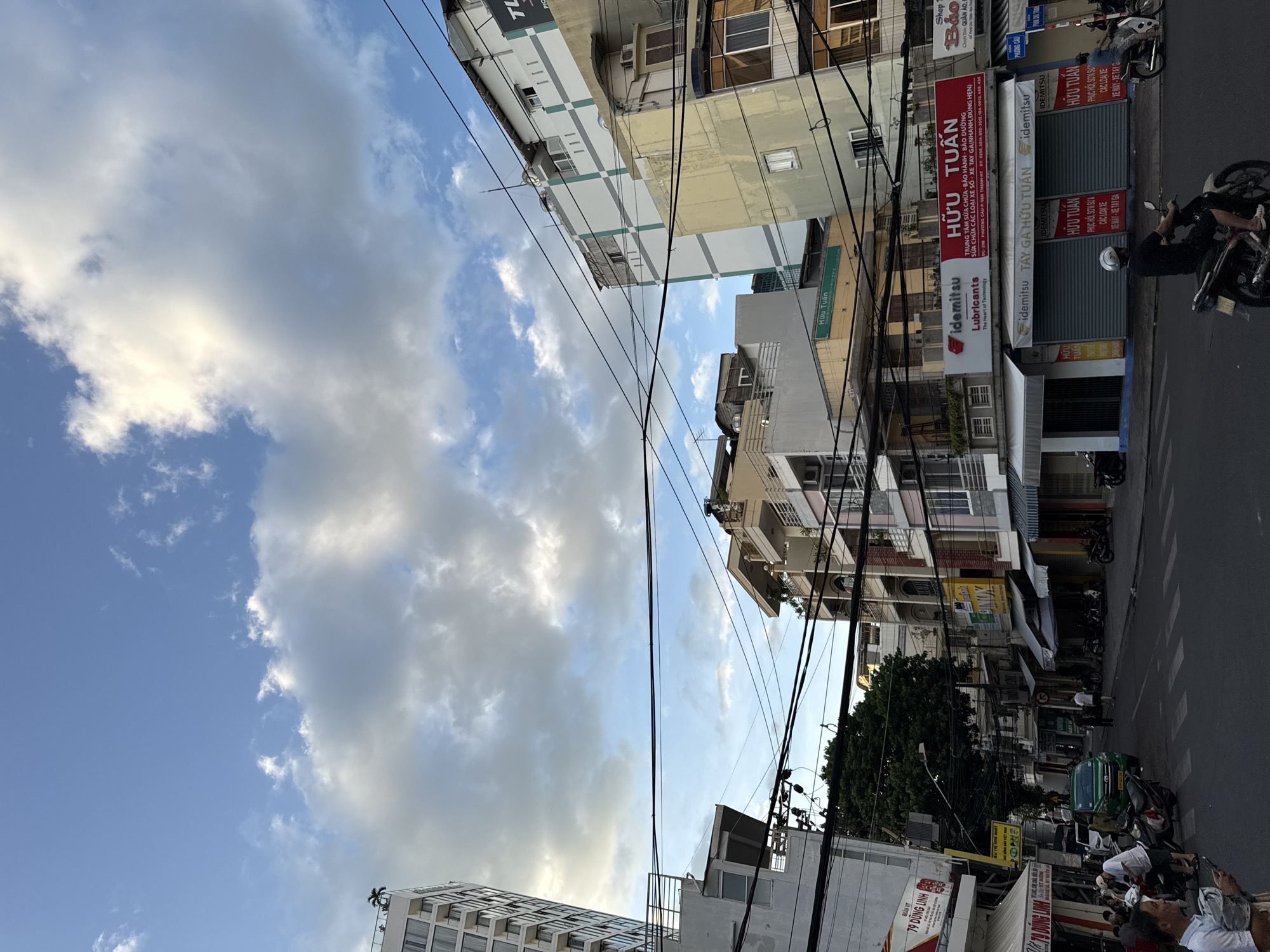 In Nha Trang, Vietnam, a street scene unfolds under a partially cloudy sky. Tall buildings line both sides of the asphalt road, adorned with numerous power lines crisscrossing overhead. On the left, a modern, multi-story building stands tall. Across the street, shops with awnings are visible, some displaying signage with Vietnamese text. A bright green car is parked on the street, and several motorcycles are present. People are seen walking and sitting on the sidewalk, and one person is riding a motorcycle. The overall atmosphere suggests a bustling urban environment.