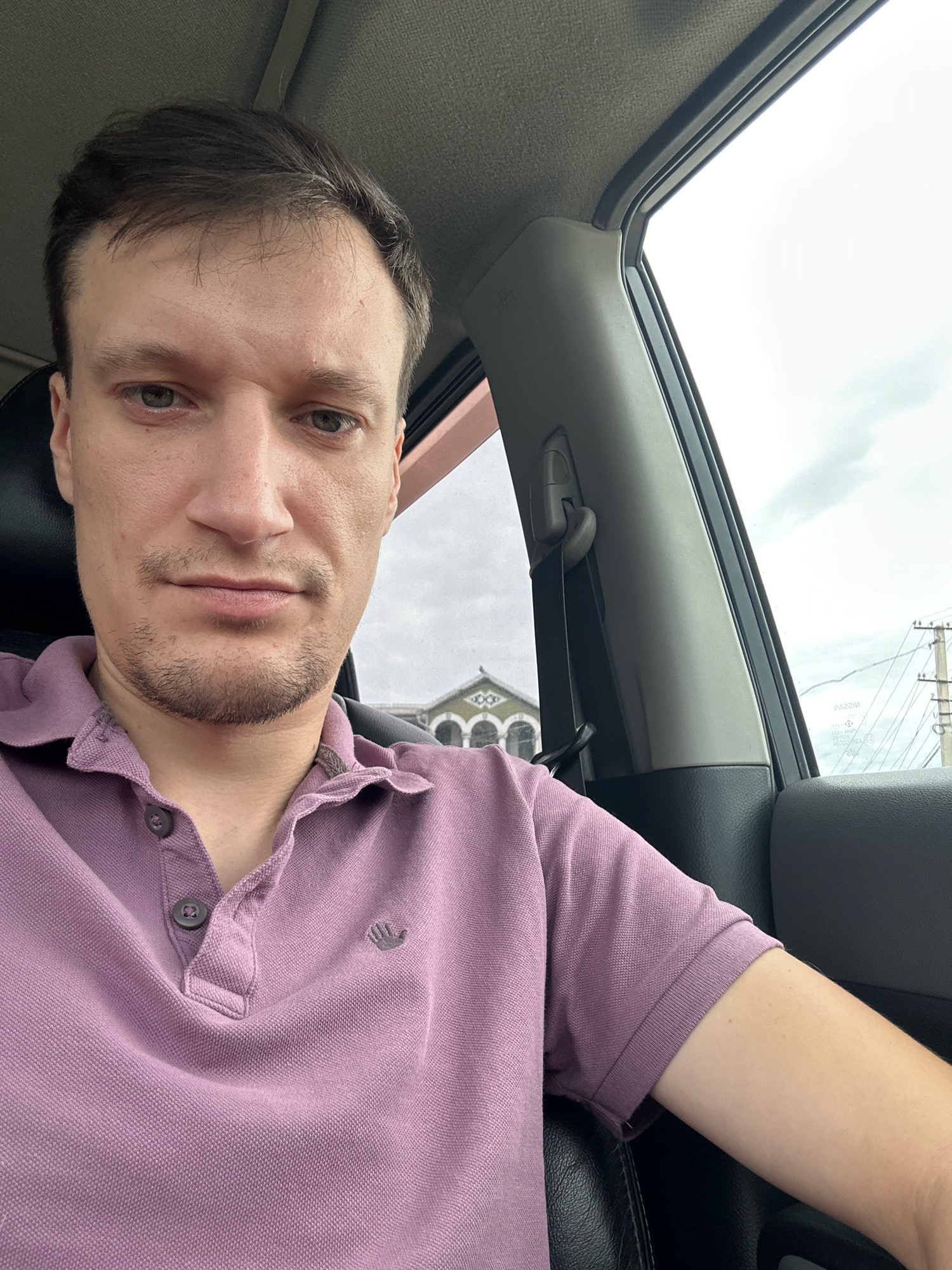 The photograph is a selfie of a man inside a car. He is wearing a purple polo shirt and has short brown hair and stubble. The car appears to be stationary. Through the car window, a building with arched windows and a cloudy sky are visible. The location is identified as Shatava, Ukraine. The image is taken from a medium-close-up perspective, focusing on the man's face and upper body.