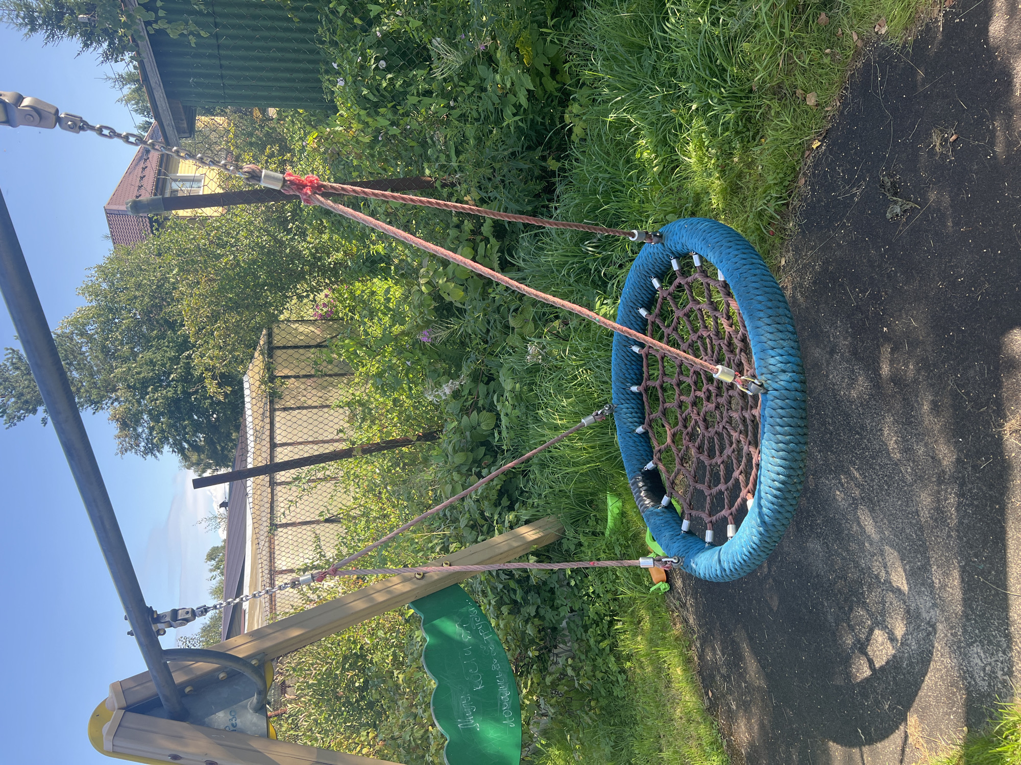 The image depicts a bird's nest swing hanging from a swing set in a yard. The swing set is constructed of wood and metal, with a weathered appearance. The swing itself is circular, with a blue rope border and a net made of brown rope. It is suspended by chains and ropes from the swing set. The background shows lush green vegetation, including trees and bushes, with a fence and a building visible in the distance. The ground beneath the swing is a dark, textured surface, possibly rubberized playground surfacing, with patches of grass and scattered leaves. Sunlight casts a shadow of the swing on the ground. A green sign with white writing is attached to the left side of the swing set. The overall scene suggests an outdoor recreational area in a residential setting, possibly a backyard or a small park. The location is specified as Роса Снт, Russia.