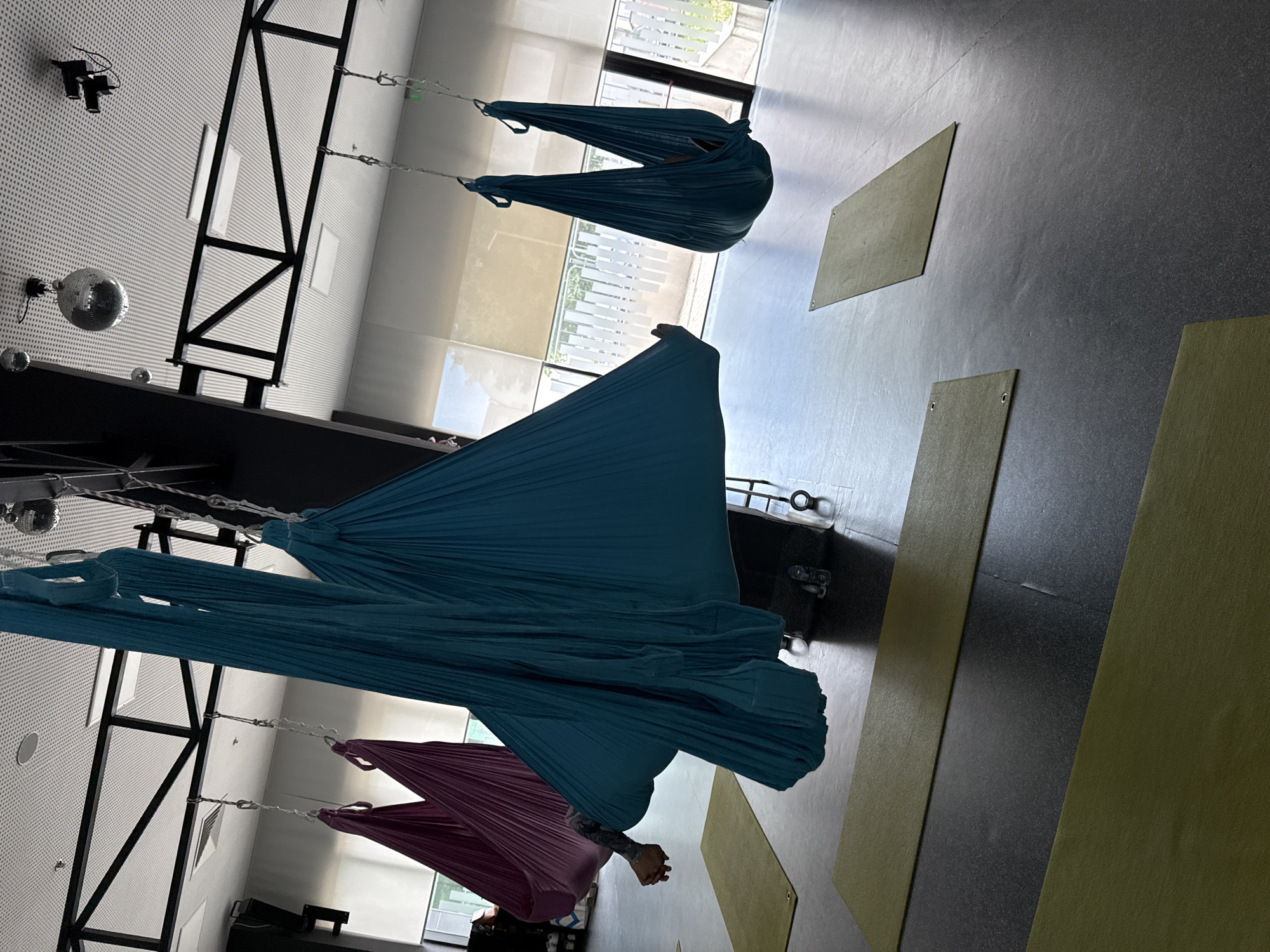 The image depicts an indoor aerial yoga studio. In the foreground, several large, colorful hammocks are suspended from the ceiling. Two large teal hammocks dominate the center of the frame, one appearing to have a person inside, indicated by an arm and part of a leg visible. To the left, two smaller purple hammocks are also visible, also appearing to be occupied. In the background, more teal hammocks are suspended. The floor is a dark, polished surface, with several yellow and olive-green yoga mats laid out. The ceiling is high and features a black metal framework supporting lighting fixtures and two disco balls. Large windows with light-colored blinds line the back wall, allowing natural light to enter the space. A black support pillar is positioned prominently in the mid-ground. The overall impression is that of a clean, modern fitness space.