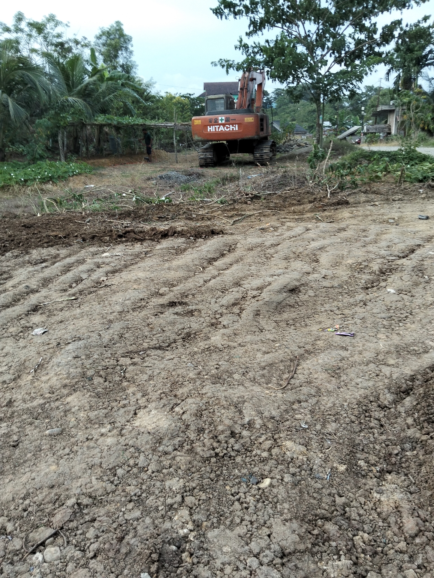 In Payagaboh, Indonesia, an orange Hitachi excavator is positioned on a cleared, dirt construction site. The ground is uneven and appears recently disturbed, with visible tire tracks from the excavator. In the background, dense green foliage and trees surround the site. A small, simple structure with a corrugated metal roof is visible to the right of the excavator. A person, dressed in dark clothing, is standing in front of a wooden structure covered in vegetation, to the left of the excavator. Several pieces of debris, including what appear to be candy wrappers, are scattered on the ground in the foreground. The sky is overcast.