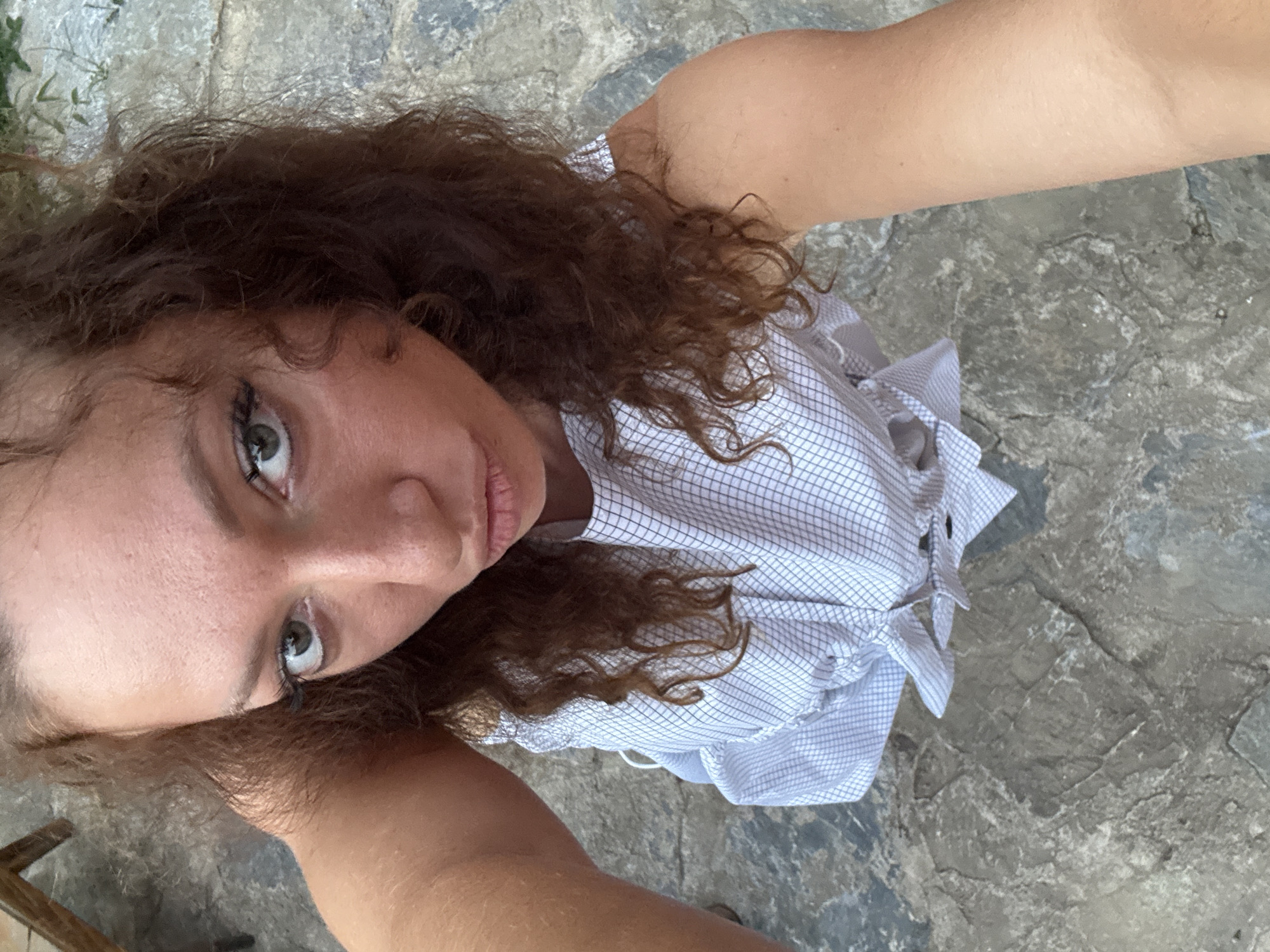 A selfie of a woman with curly brown hair and green eyes wearing a light blue and white checkered shirt. The background is a stone surface, potentially a pavement or courtyard. The image was likely taken outdoors in daylight. No specific identifiers indicate the location as Bắc Hà, Vietnam.