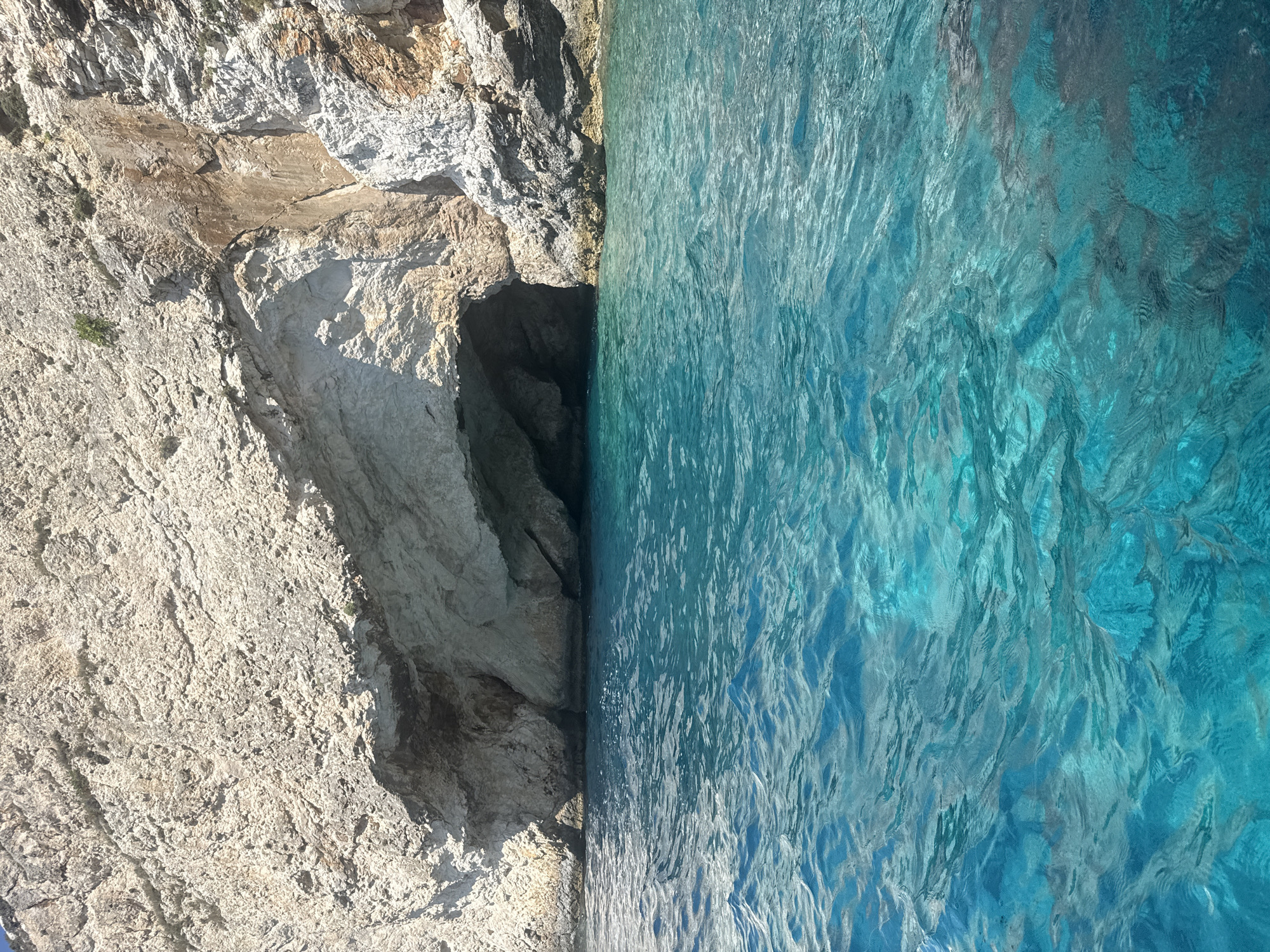 A view of a sea cave entrance along a rocky cliff face, with clear turquoise water in the foreground. The cave is dark and recessed into the light-colored rock. The water exhibits ripples and reflections of the sunlight, with some submerged rocks or seabed visible in the clearer sections. The cliff face is rough and textured, with areas of lighter and darker rock. There are no people or man-made objects visible in the image.