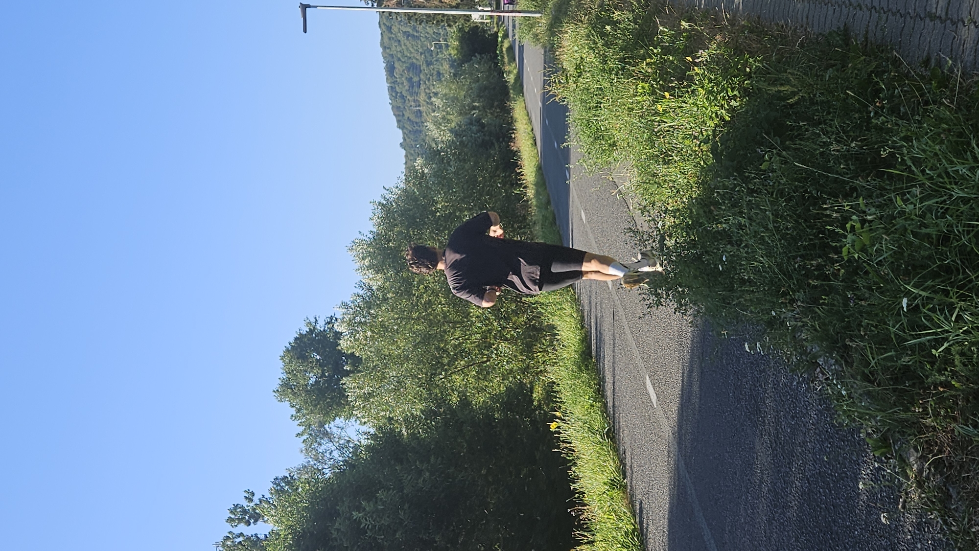 An individual is running on a paved path. The runner is seen from behind, wearing a dark t-shirt and dark shorts, with white socks and light-colored running shoes. The path is bordered by green grass and vegetation on both sides. In the background, there are trees and a hill. The sky is clear and blue. The scene appears to be outdoors, possibly in a park or on a trail within or on the outskirts of Prešov, Slovakia, given the natural landscape.