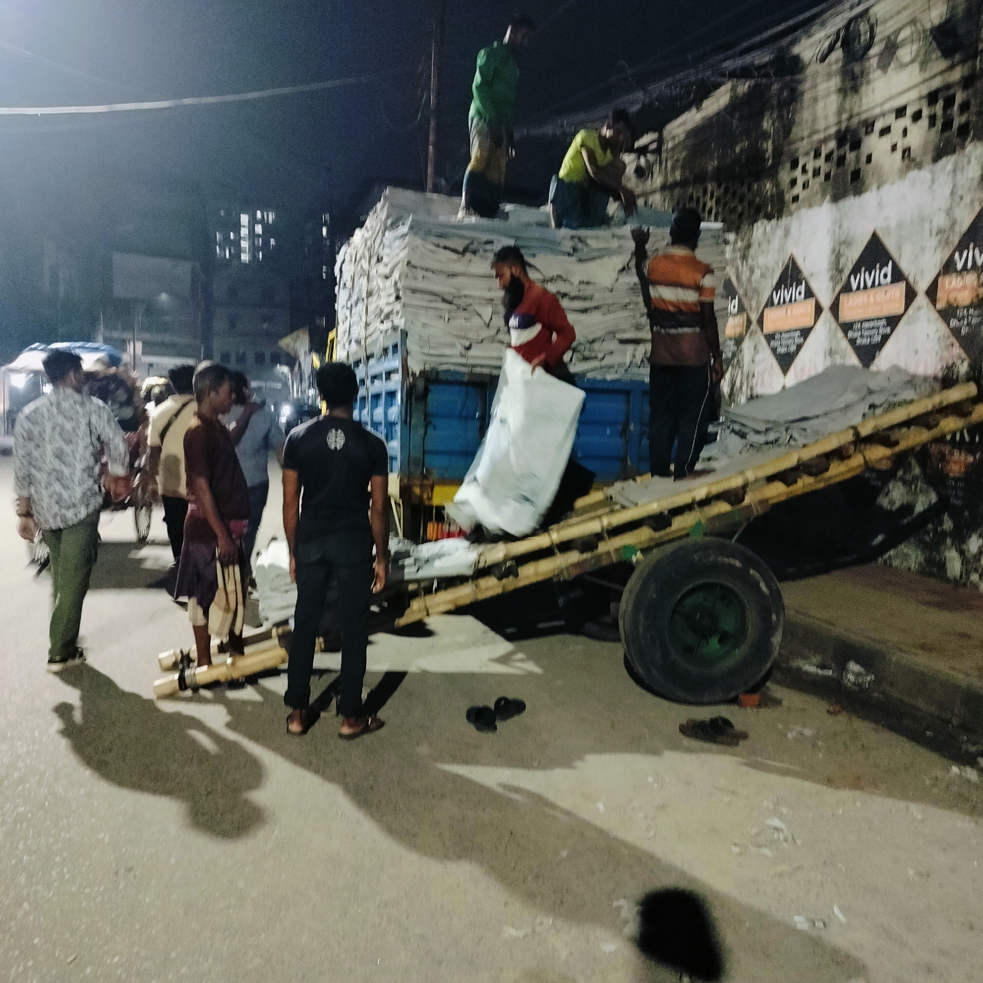 The image captures an active street scene at night in an urban area of Dhaka, Bangladesh, depicting multiple men engaged in the manual loading or unloading of goods. A large blue truck or heavy-duty cart, piled high with numerous white, rectangular packages or sacks, is central to the activity. A makeshift ramp constructed from bamboo poles extends from the ground to the back of the vehicle, serving as an incline for moving the packages.
At least nine men are visible, some actively working and others observing. Two men are standing on top of the stacked packages on the vehicle. Another man, wearing a red and white shirt, is seen guiding a package down the bamboo ramp. Other men are positioned on the street, assisting with the packages or standing nearby. The scene is illuminated by artificial light sources, casting long, distinct shadows of the figures and objects onto the street.
To the right, a building wall with a rough, textured surface and some foliage or grime is visible. Several black, diamond-shaped posters are affixed to this wall. These posters clearly display the brand name "vivid" and text below it that reads "LADIES & GENTS." Further legible text includes "1/A Naya Paltan, Dhaka 1209, Bangladesh," providing a specific location cue. Footwear, including sandals and slippers, can be seen scattered on the ground near the activity. The image conveys a sense of industrious manual labor taking place during the late hours in a bustling city.