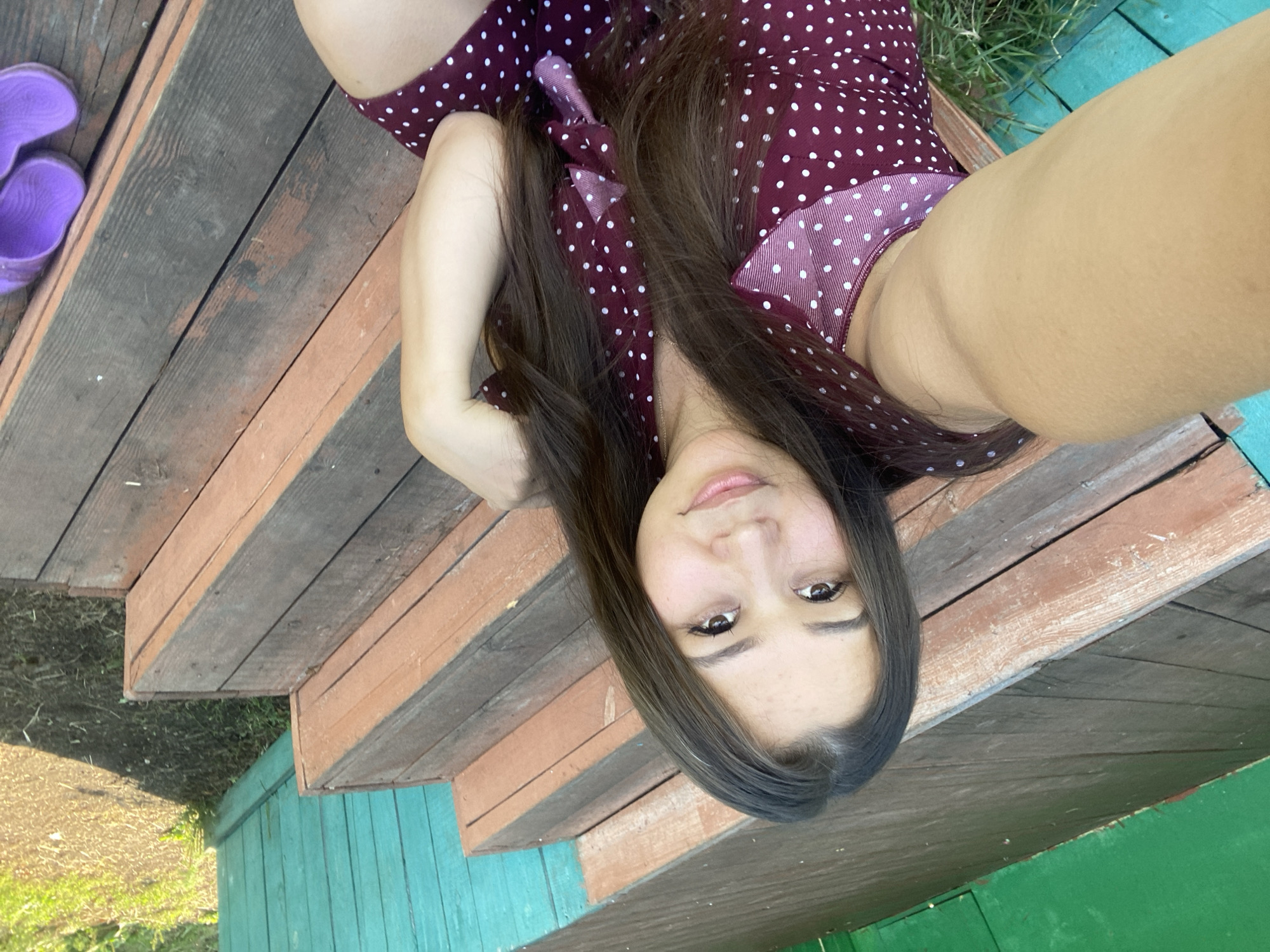 A young woman with long, dark hair is taking a selfie while sitting on wooden steps. She is wearing a dark red dress with white polka dots. The steps appear to be outdoors, possibly on a porch or deck, with a green and turquoise wooden fence in the background. Sunlight casts shadows on the ground and steps. A pair of purple sandals are visible on the far right of the frame. The scene is likely in Maloarslangulovo, Russia, given the provided context.