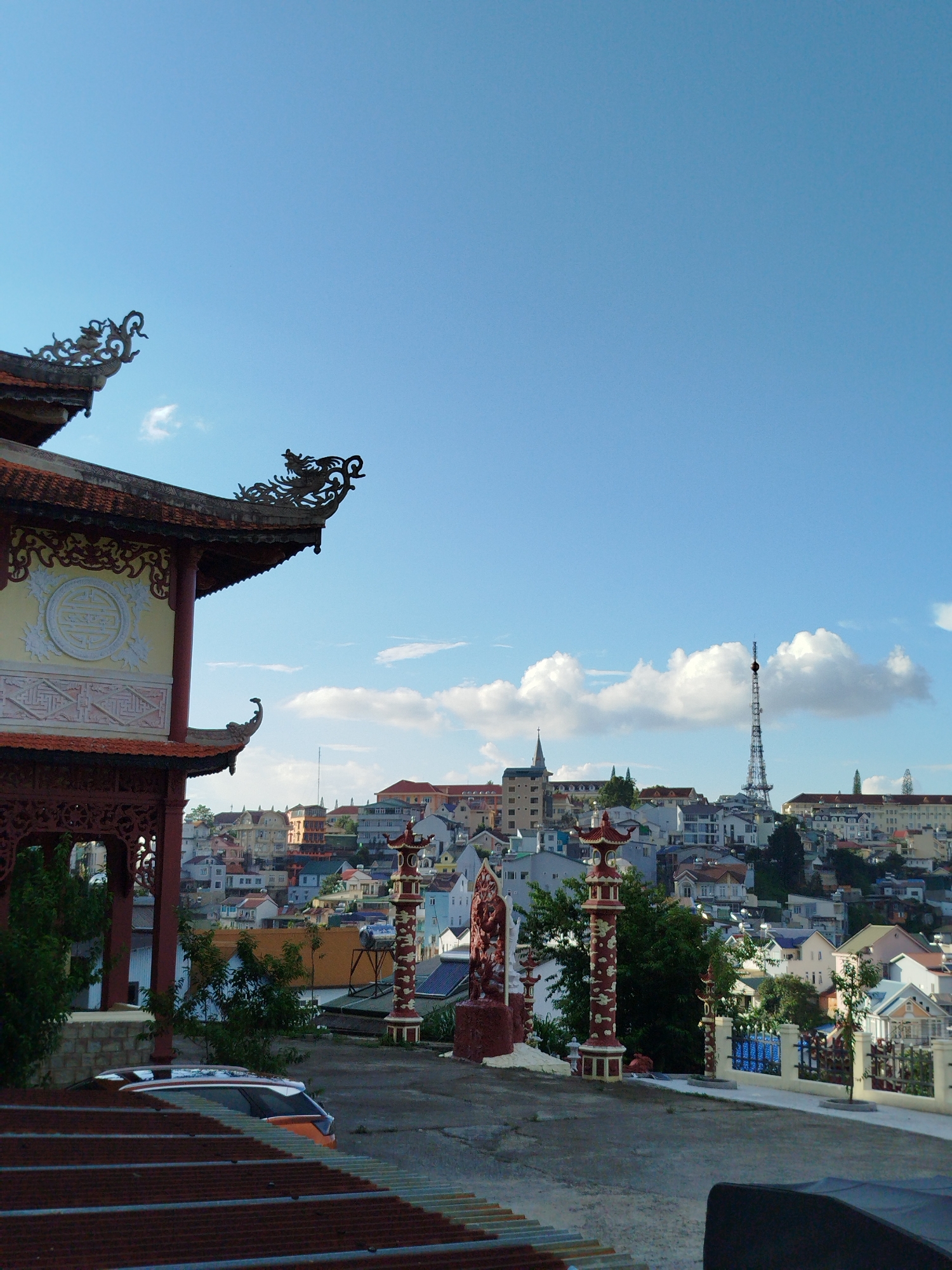 The image captures a scenic view of Da Lat, Vietnam, with a traditional pagoda in the foreground overlooking a densely built city. The pagoda's ornate architecture, characterized by intricate dragon carvings and red and gold accents, dominates the left side of the frame. A vast expanse of blue sky with scattered white clouds stretches above. In the mid-ground, a cluster of colorful buildings with varying architectural styles, typical of Da Lat, cascade down a hillside. A prominent transmission tower stands tall in the distance, piercing the skyline. The foreground shows a paved courtyard with decorative pillars, a statue, and a car. The overall impression is one of a charming, hilly city bathed in natural light.