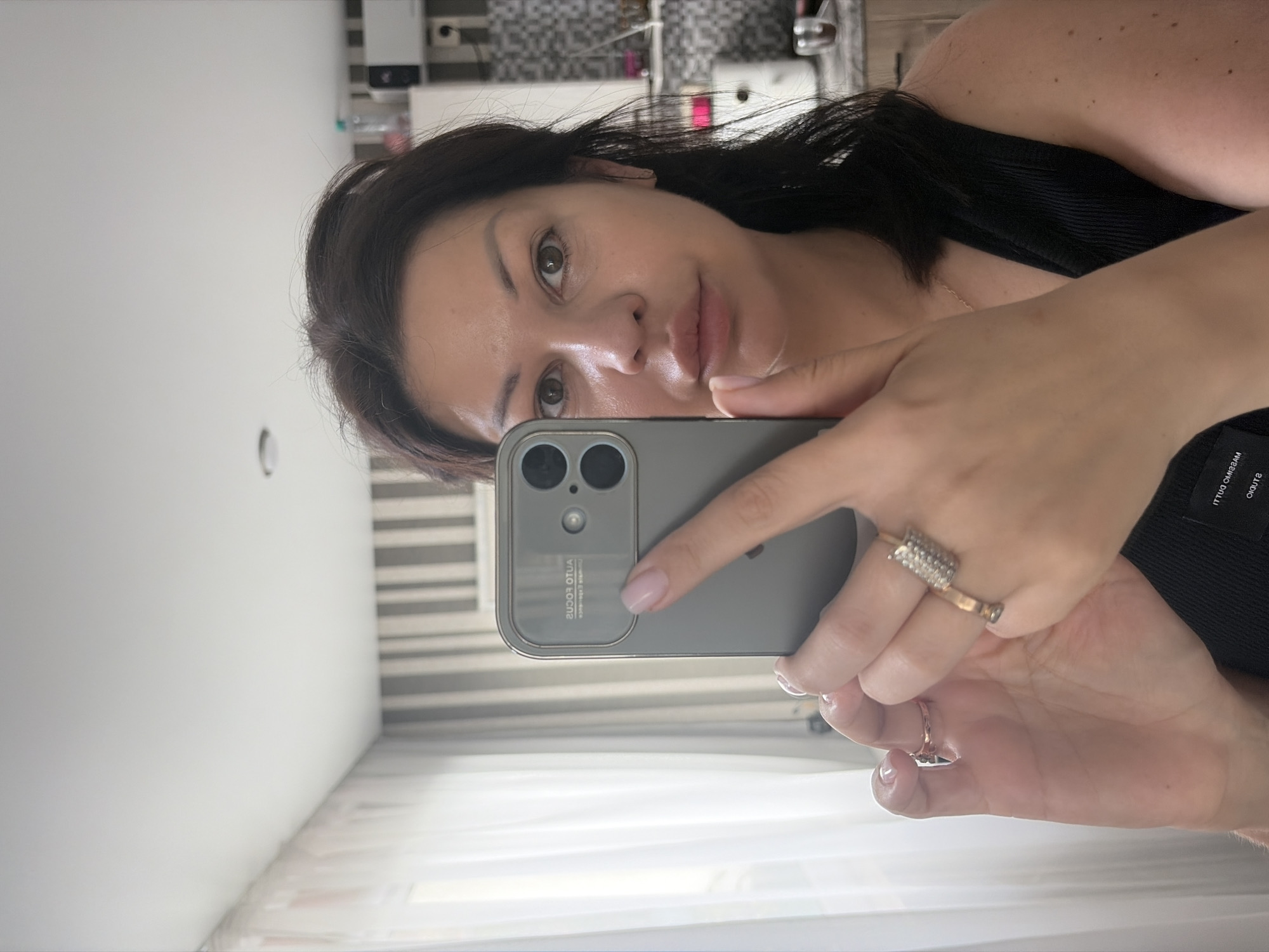 A woman is taking a selfie in a room in Sochi, Russia. She is holding a gray smartphone in her right hand, positioned to capture her reflection in a mirror. The woman has dark hair, brown eyes, and fair skin. She is wearing a black tank top and several rings on her fingers. The background shows a wall with vertical stripes, a window with sheer curtains, and a kitchen counter with some appliances. The lighting is natural, coming from the window.