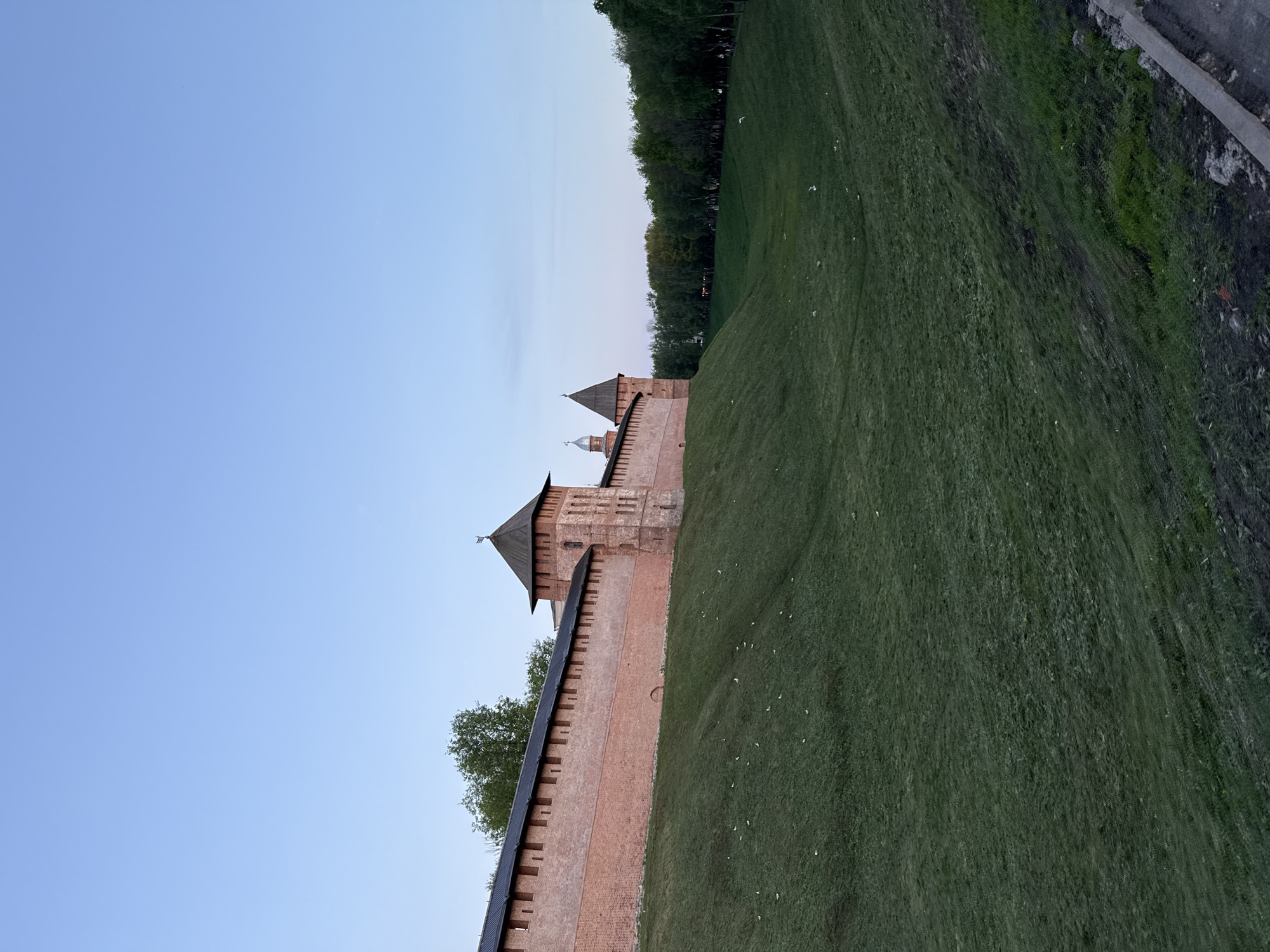 This landscape photo depicts a section of the Veliky Novgorod Kremlin walls, Russia, captured at an oblique angle. The walls, constructed of red brick and featuring battlements, stretch along a grassy embankment on the right side of the image. Two prominent towers with dark pointed roofs punctuate the wall’s design, with a smaller dome-topped structure visible behind them. The sky is a gradient of light blue, suggesting either dawn or dusk. A line of trees runs parallel to the embankment, providing a backdrop. The grass is cut short and sparsely populated with small white objects, possibly birds. The absence of people, other structures, or vehicles indicates the setting is focused solely on the historic architecture within a natural context.
