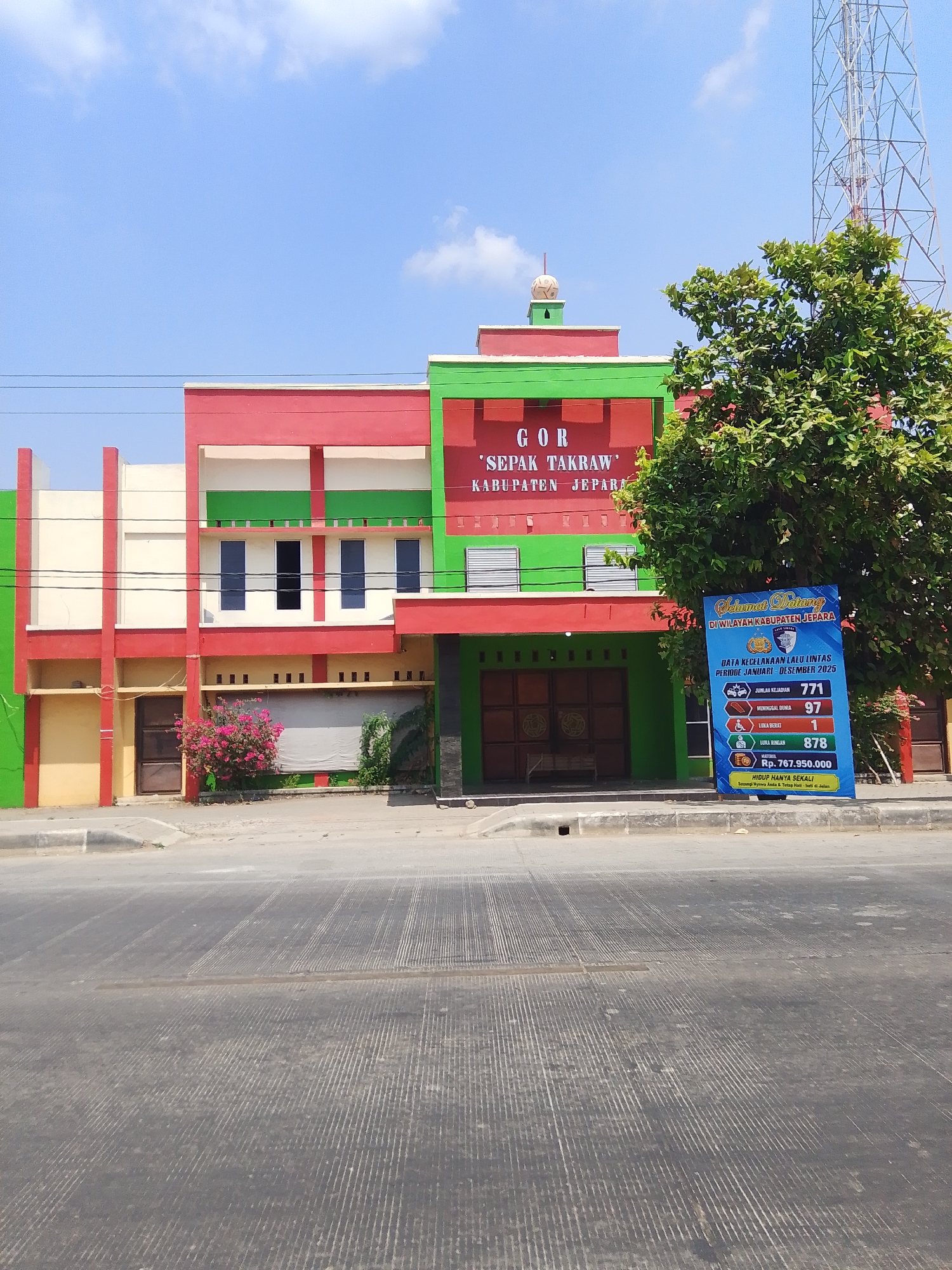 The image captures a street view of a building with a sign that reads "GOR 'SEPAK TAKRAW' KABUPATEN JEPARA". The building has a striking red and green facade. To the right of the building, a tall communication tower is visible, partially obscured by a large tree with green foliage. In front of the building, on the sidewalk, a blue banner displays traffic accident data for the Jepara Regency from January to December 2025. The banner details the number of incidents, fatalities, injuries, and material losses, along with a slogan promoting road safety. The street in the foreground is asphalt with distinct grooved markings. The sky is clear blue with scattered white clouds. The overall scene suggests a public facility located in Jepara Regency, Indonesia, with an emphasis on safety and sports.
