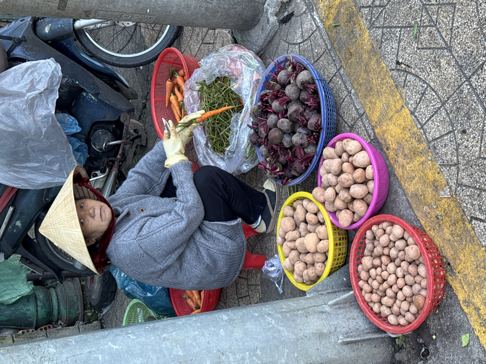 A person wearing a conical hat and a grey sweater is squatting on a red stool next to a motorcycle on a sidewalk in Da Lat, Vietnam. They are wearing yellow gloves and holding a knife and a carrot, appearing to peel it. In front of the person are several baskets of produce: a red basket with carrots, a blue basket with beets, a yellow basket with potatoes, a pink basket with potatoes, and a red basket with more potatoes. The sidewalk is paved with gray and white tiles, and a yellow line runs across it. There is a large green pillar on the left side of the image.