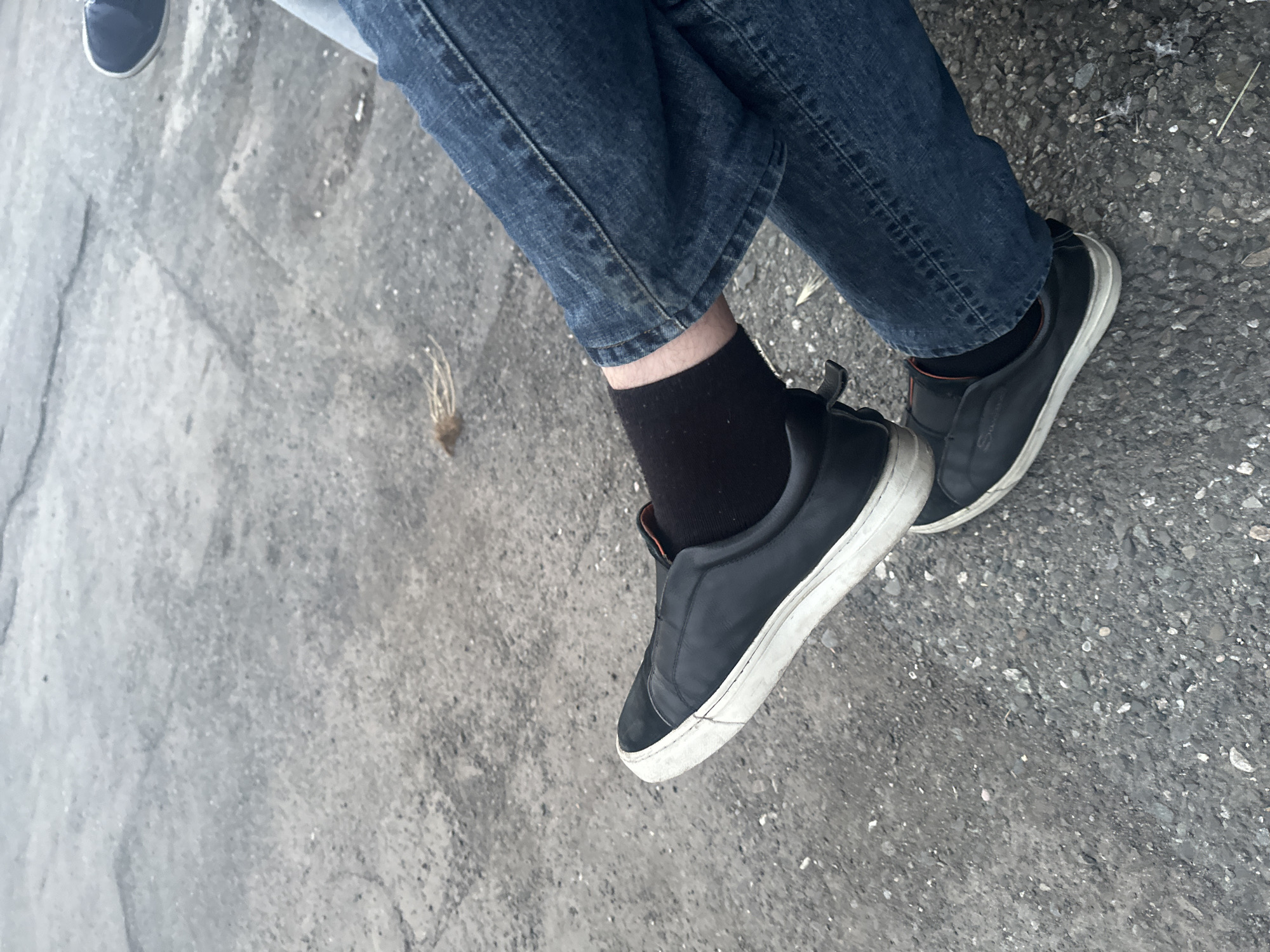 The image shows a person wearing blue jeans, black socks, and black slip-on shoes with white soles. The person is sitting on a concrete surface that appears to be a sidewalk or pavement. The ground has a rough texture and some minor cracks. There is a small clump of dead grass or weeds nearby. Based on the location, the image is likely from Yerevan, Armenia. The photo composition focuses on the lower portion of the body.