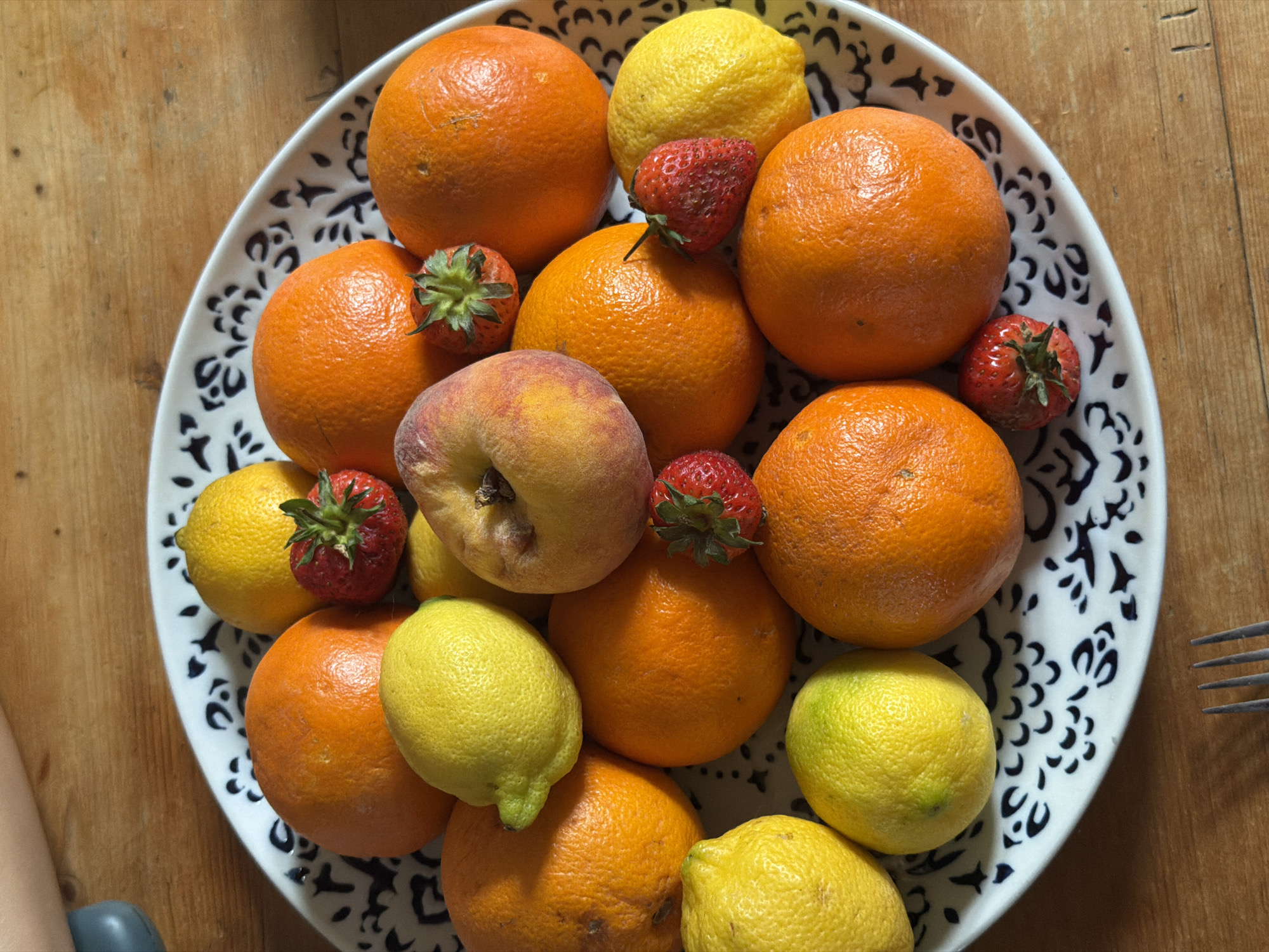 A top-down shot shows a white plate with a black floral pattern filled with fruit. There are oranges, lemons, strawberries, and a peach. The plate sits on a wooden surface. A fork is visible on the right side of the plate. No people are fully visible, but part of an arm is in the bottom left corner. The location is unknown and no location information is apparent.