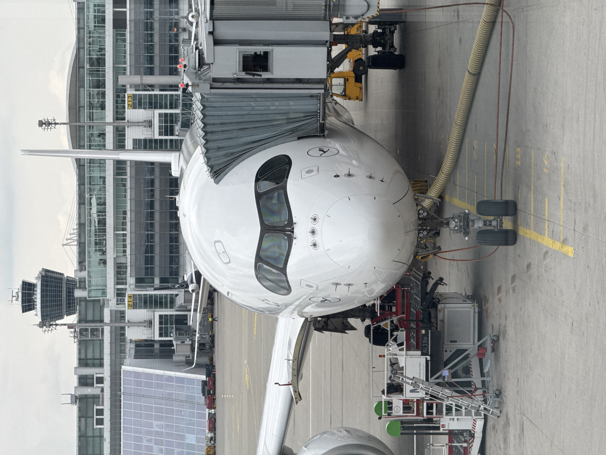 The image shows the nose of a Lufthansa Airbus A350 aircraft connected to a jet bridge at Munich Airport (Flughafen München), located in Oberding, Germany. Airport ground support equipment is present around the aircraft's nose and front landing gear. In the background, the airport terminal building is visible with numbered gates. The scene is overcast, suggesting daylight. No people are visible in the frame.