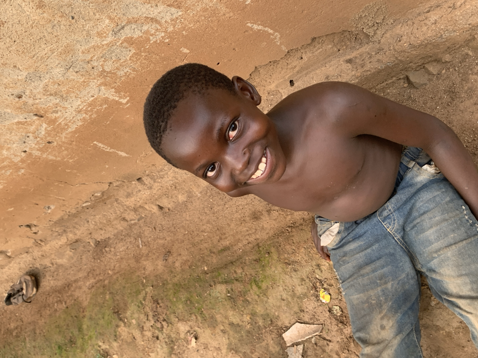 A young, dark-skinned boy stands smiling in what appears to be a dirt-covered courtyard or yard. He is shirtless, wearing only blue jeans. The background includes a tan, textured wall or surface on the left, and bare dirt and debris on the ground. There is a bag and a broken piece of ceramic tile on the ground. The environment suggests a lower-income area in Agban, Nigeria.