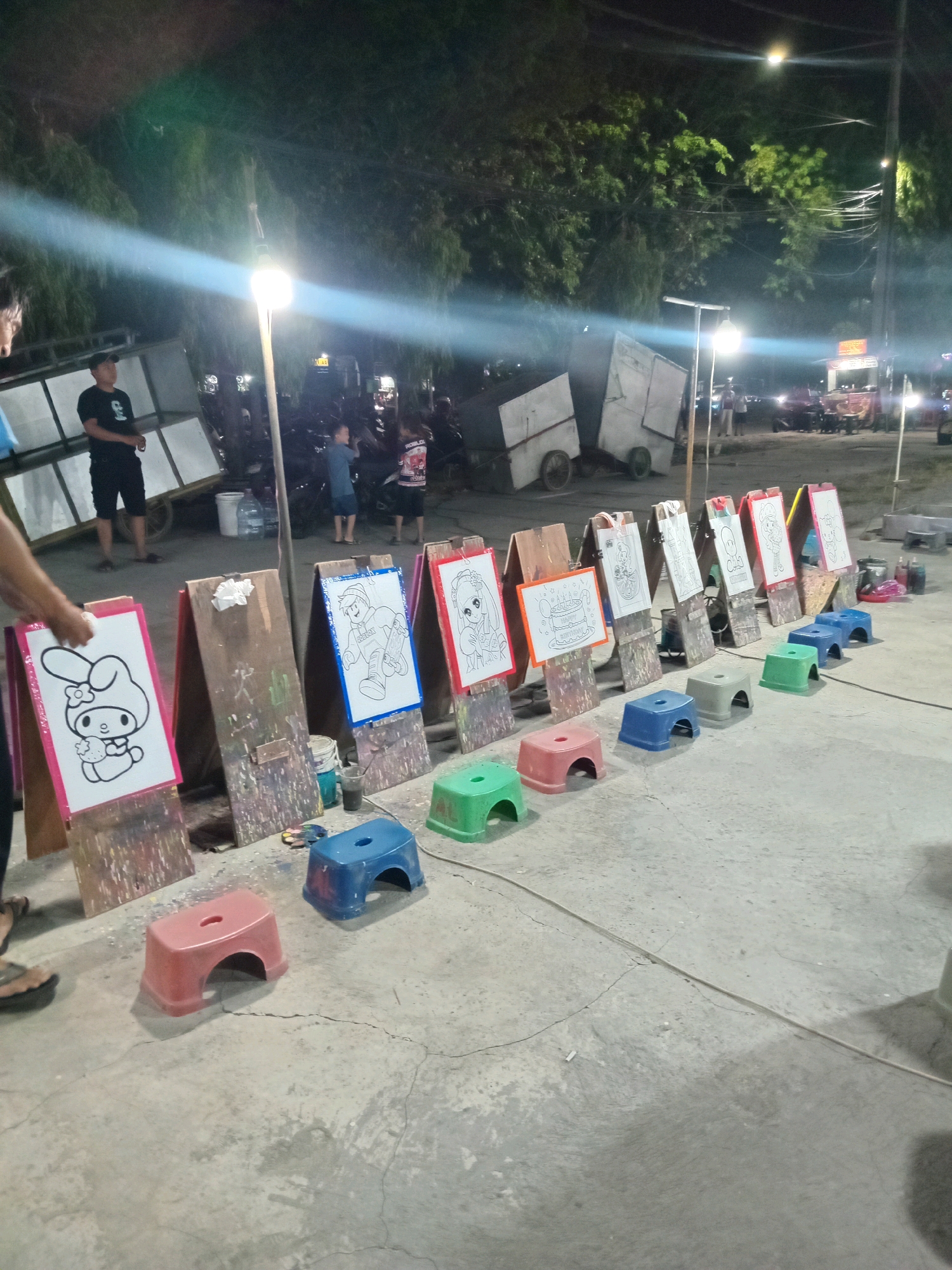 The image shows a nighttime scene in Sedati Agung, Indonesia, likely at a street market or outdoor event. In the foreground, a row of easels displays children's coloring pages. Colorful plastic stools are placed in front of the easels, suggesting a children's activity area. In the background, several people are visible, including adults and children, standing around and observing the scene. Several carts and vehicles are also present, hinting at a commercial or entertainment setup. The environment appears to be an urban outdoor space with trees and streetlights illuminating the area.