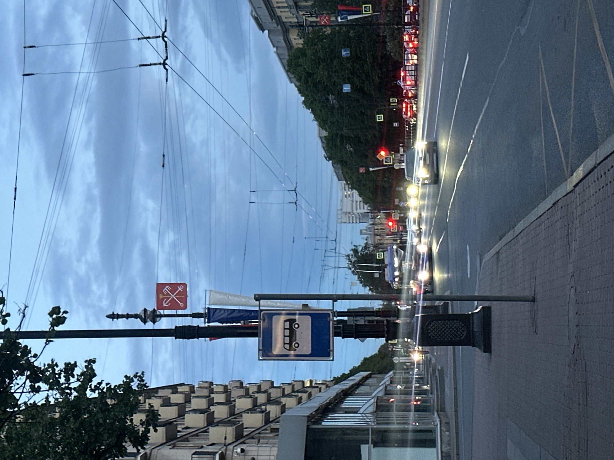 A street scene at dusk, likely in an urban area, possibly St. Petersburg, Russia, given the presence of trams and architectural style. The sky is overcast with a blue-grey hue. A multi-story building with balconies lines the left side of the street. A sidewalk is paved with bricks, and a lamppost with a decorative base stands on the sidewalk, displaying a bus stop sign. The street itself has multiple lanes with vehicles in motion, their headlights creating streaks of light due to motion blur. A bus stop sign with a pictogram of a bus is visible. Other traffic signs and banners are also present on lampposts. Trees are visible along the right side of the street. Electrical wires crisscross overhead.