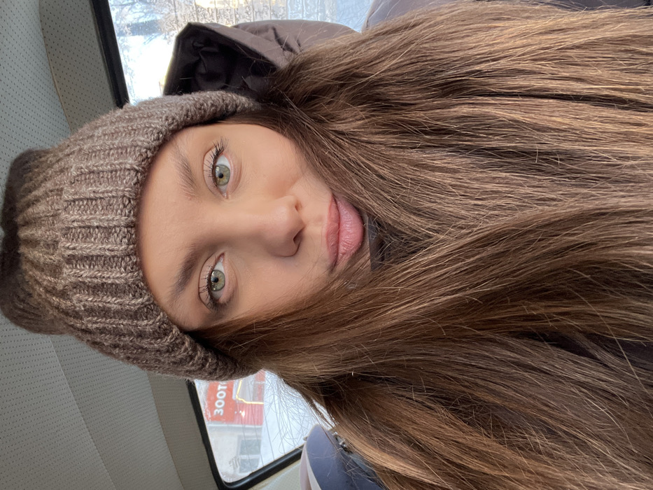 A young woman with light green eyes and long, light brown hair wears a ribbed brown knit hat and a dark brown hooded jacket. She appears to be inside a vehicle, as suggested by the window and textured interior wall. A red and white sign is visible through the window. Her lips are slightly parted.