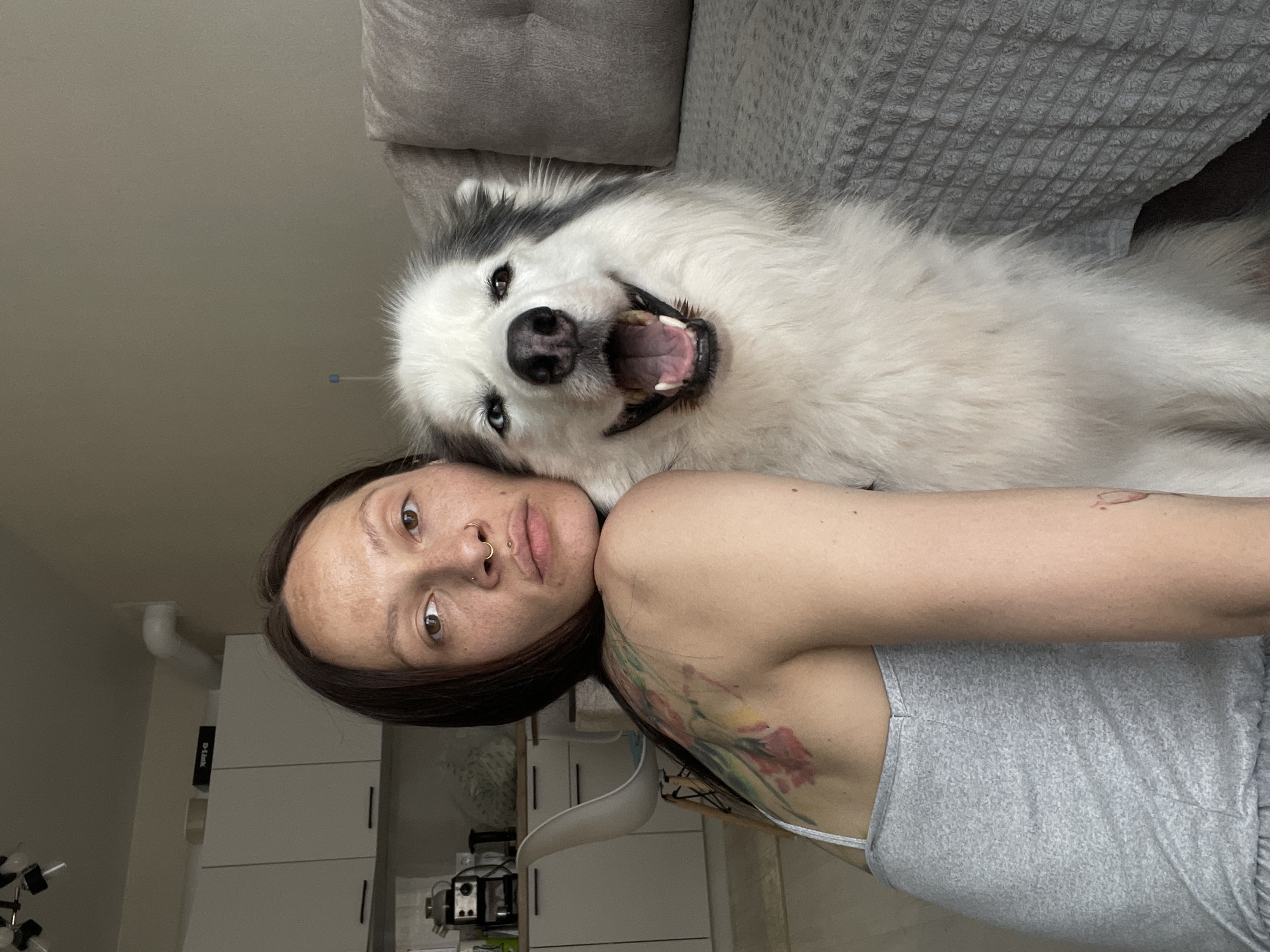 A woman lies on a grey couch with her head and upper body visible in the foreground. She has brown hair, freckles, a nose ring, and a floral tattoo on her shoulder. She is wearing a grey tank top. A large white and grey dog lies on the couch with its head resting on the woman's head. The dog is smiling, exposing its teeth. In the background, there is a white cabinet, a floor lamp, and a coffee maker. The image provides no information about the location in the city of Pervomaysky, Russia.