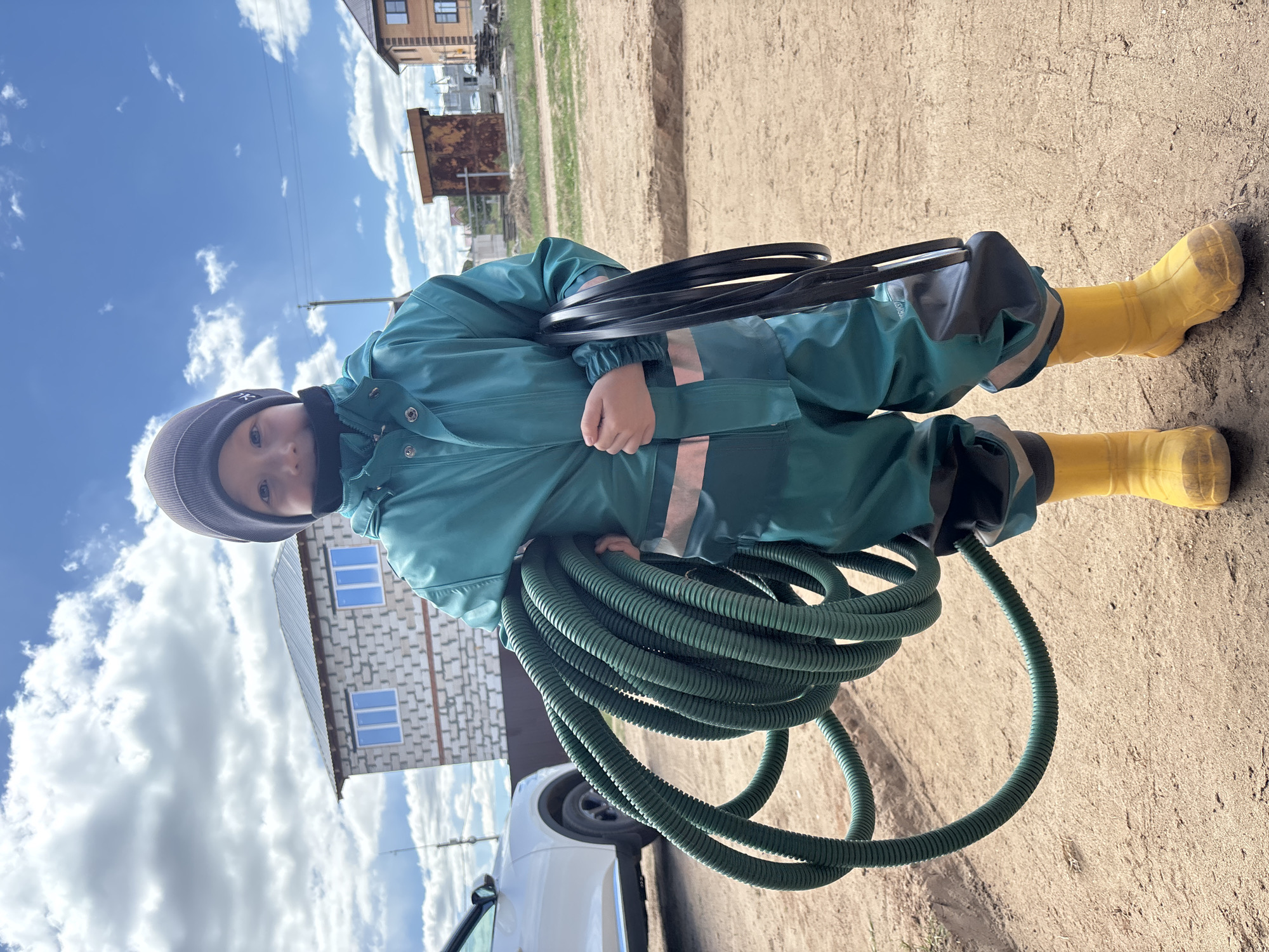 A child is centered in the frame, wearing a teal-colored jumpsuit with reflective stripes and yellow rubber boots. The child is also wearing a gray knit hat and a black face mask. They are holding a coil of green, corrugated hose and a black rubber seal.
The background includes elements of a suburban environment. A white car is partially visible on the left. Further back, a building with blue-framed windows is seen. In the far background, there are other buildings and vegetation. The ground appears to be a dirt or sandy surface. The sky is visible with scattered clouds.
The location may be in the city of Chaikovsky, Russia.