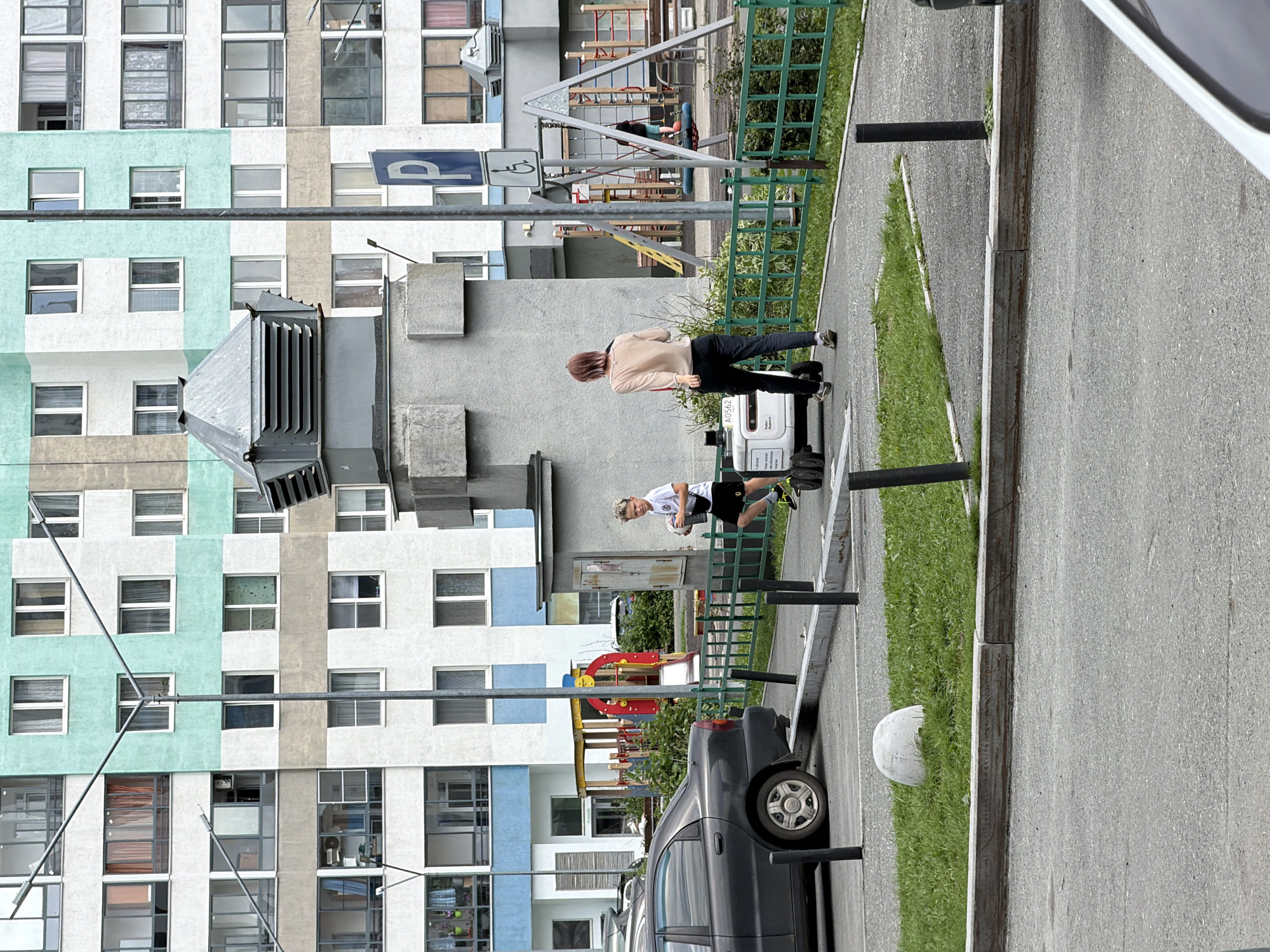 An outdoor daytime shot shows a residential building with a playground and a parking lot. The building, painted in alternating horizontal bands of white, teal, and beige, has numerous windows and balconies. A silver-gray car is parked in a lot in front of the building, to the left of the frame. A green metal fence separates the parking lot from a paved walkway where a white delivery robot is moving. A young boy in a white t-shirt and black shorts walks beside the robot, holding a white object. A woman with short pinkish-brown hair, wearing a light-colored top and dark pants, walks in front of the robot, facing away from the camera. Behind the fence, to the right, is a playground with swings and slides. A blue parking sign with a white "P" and a wheelchair accessibility symbol is visible mounted on a pole near the playground. The ground is paved with asphalt and concrete, with patches of grass. This scene is consistent with the architecture and urban planning typical of residential areas in Yekaterinburg, Russia.