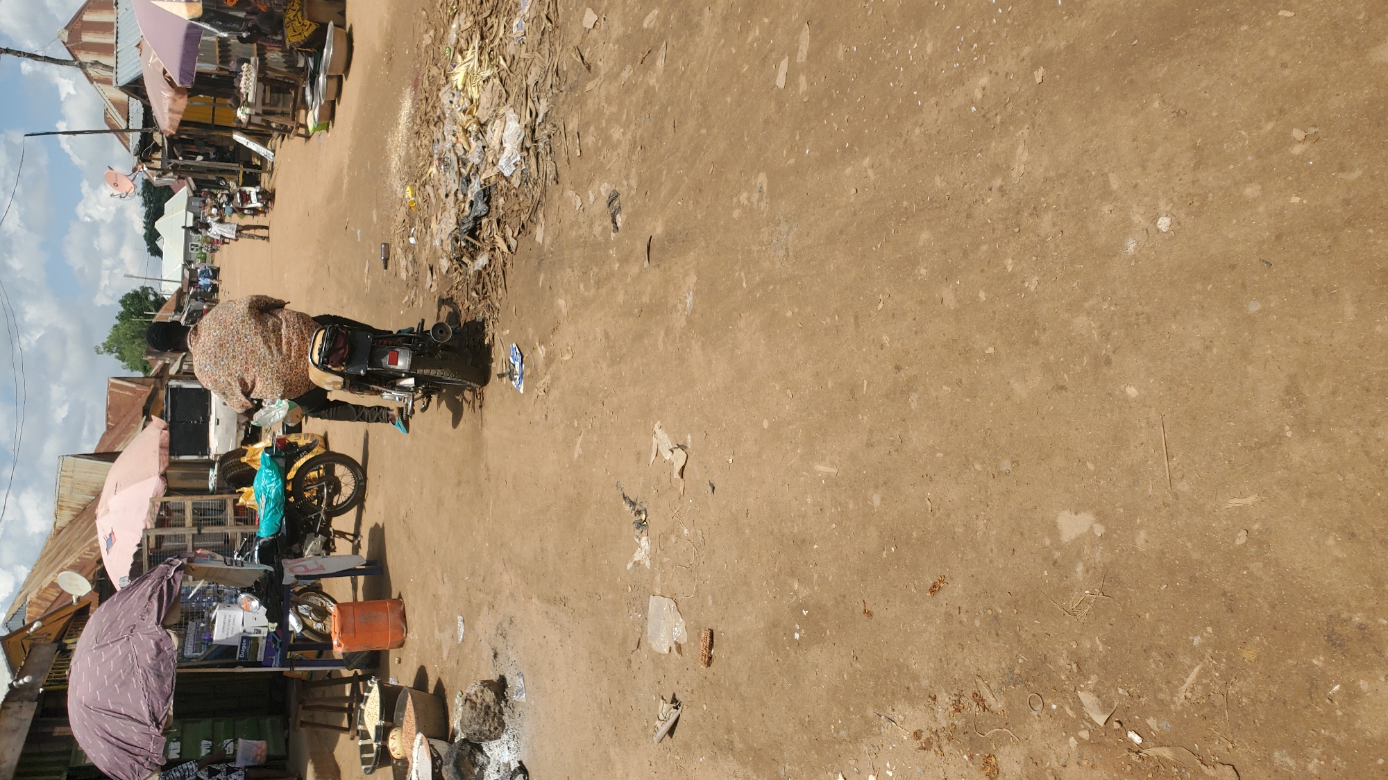 The image depicts a busy marketplace scene in Jalingo, Nigeria. A person is riding a motorcycle down a dirt road lined with market stalls. The stalls have corrugated metal roofs and display various goods, including food items like grains and produce. Umbrellas provide shade over some of the stalls. The ground is dusty and littered with debris. Other people and vehicles are visible in the background, suggesting ongoing commercial activity.
