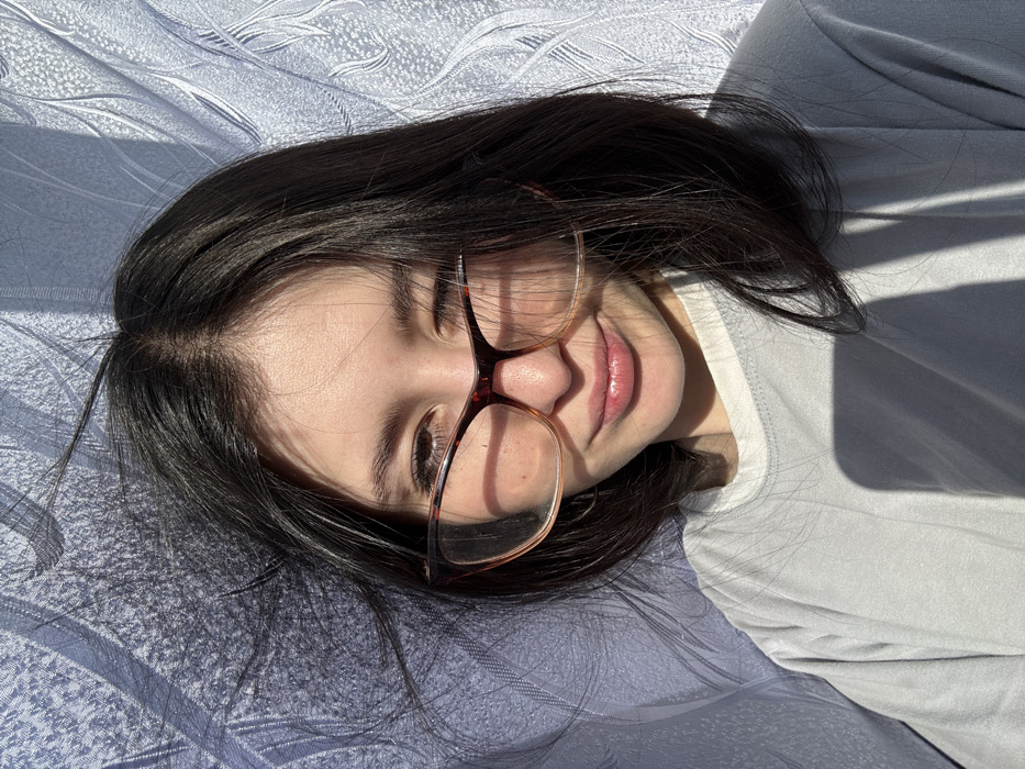 A portrait features a young female individual with dark brown, shoulder-length hair and fair skin, positioned against a textured, light-colored fabric background, likely a curtain, displaying an undulating pattern. She is wearing eyeglasses with tortoiseshell-patterned frames, transitioning from dark to light. Her right eye is open, while her left eye is partially closed, and her lips form a subtle smile. Direct, strong natural light originates from the viewer's left, casting pronounced shadows across her face, particularly on her left cheek and neck, and onto her light grey t-shirt with a white collar band. The light also creates highlights and shadows on the background fabric, emphasizing its texture. The individual's hair appears slightly dishevelled on the left side. The scene is located in the city of Фоки, Russia.