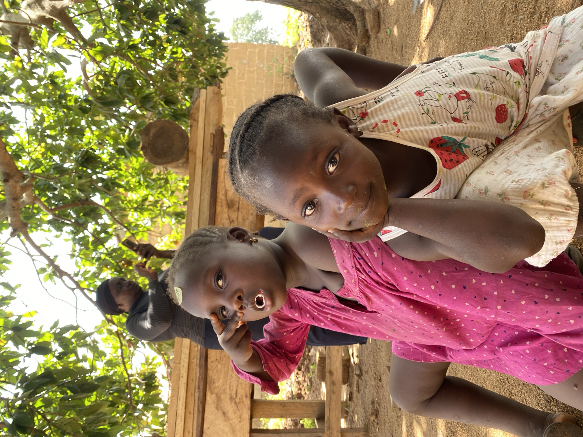 Two young dark-skinned girls are the central subjects of the image. One wears a pink dress with small white hearts, and the other wears a white tank top with strawberry prints. An adult stands in the background with a hijab and is holding a mobile device. The setting appears to be an outdoor area, possibly Hoss, Nigeria, given the location specified. The scene is bathed in sunlight. The environment consists of a makeshift wooden structure, trees with dense foliage, and what appears to be a brick wall. The foreground is sandy and covered in loose dirt.