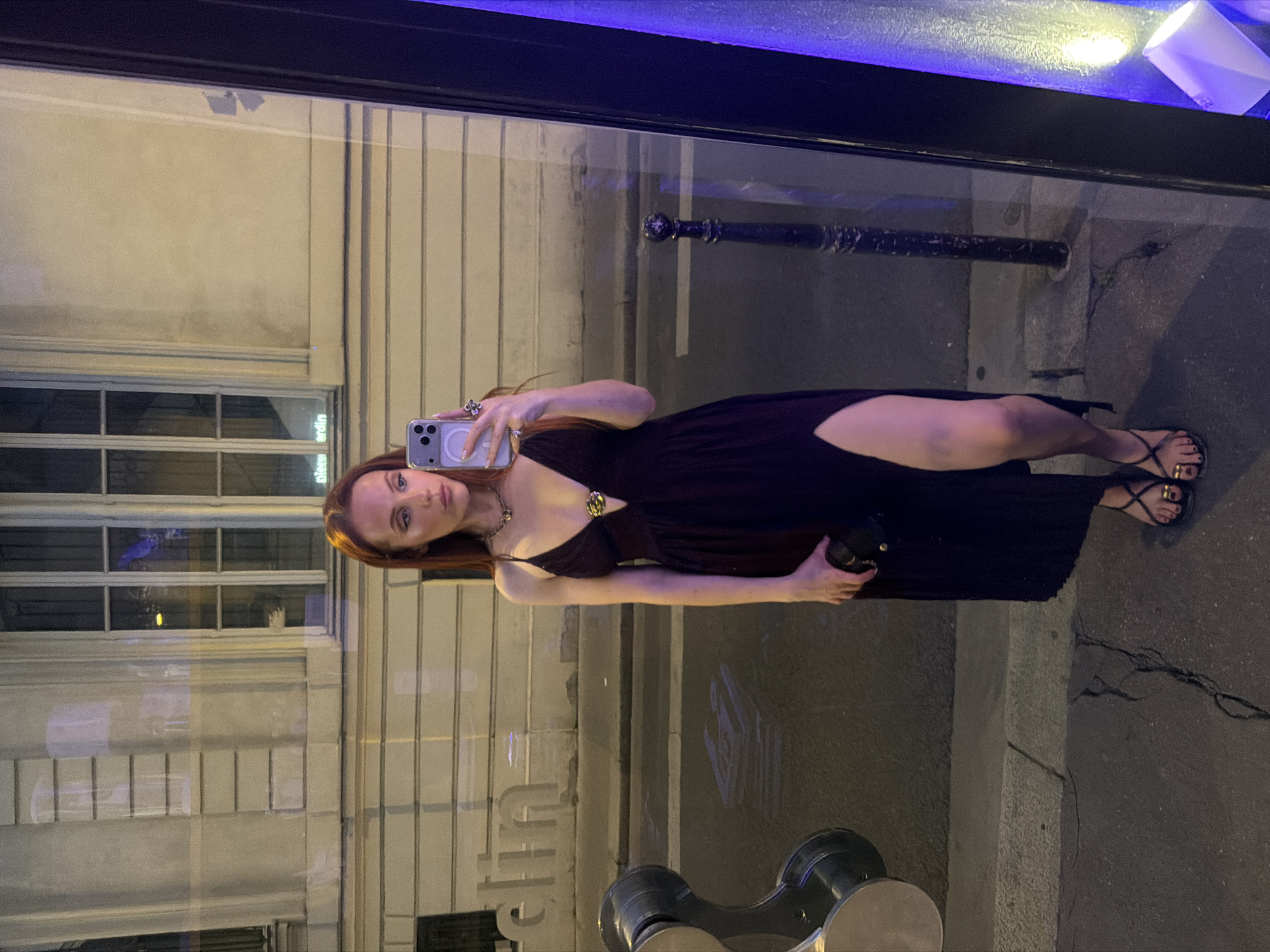 A woman with long red hair takes a selfie in front of a shop window at night. She wears a dark dress with a thigh-high slit and black sandals. The woman holds a black clutch in her left hand and a phone in her right. The name "Berlin" is reflected in the shop window. The surrounding environment features a building with numerous windows and a Parisian street with bollards. The photograph likely captures a moment in Paris, France.