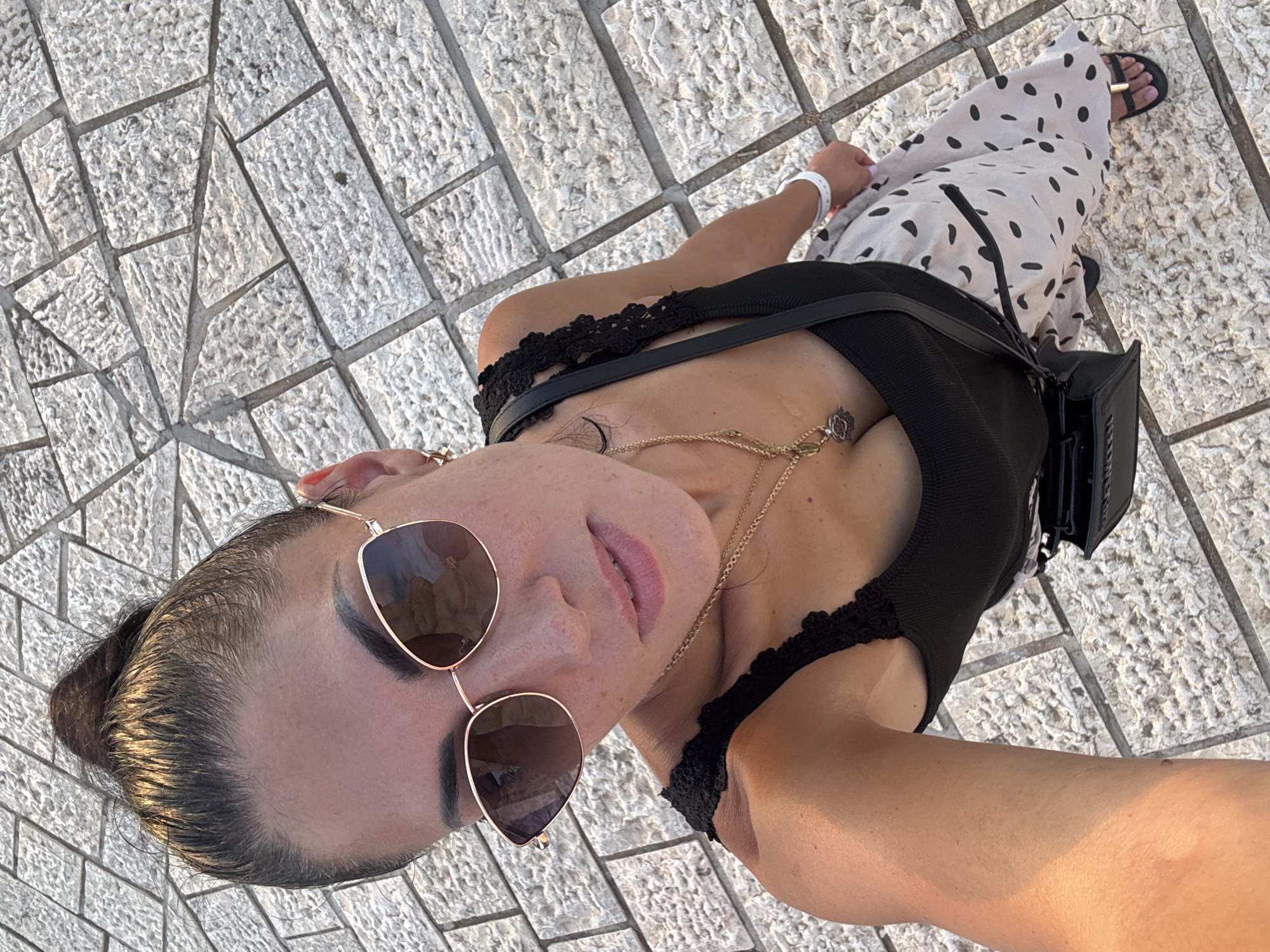 A woman is captured in a high-angle, selfie-style shot, standing on a paved walkway composed of large, light-colored rectangular stones arranged in a radiating pattern. She is wearing a black tank top with lace trim, a light-colored skirt with black polka dots, sunglasses with rose gold frames, and black sandals. A black crossbody bag with a gold buckle hangs from her shoulder. She has her dark hair pulled up in a bun and is wearing gold necklaces, one with a circular pendant. A white wristband is visible on her left wrist. The scene is outdoors, with the paving stones suggesting a public area like a promenade or plaza, potentially in Tel Aviv, Israel, given the architectural style of the paving.