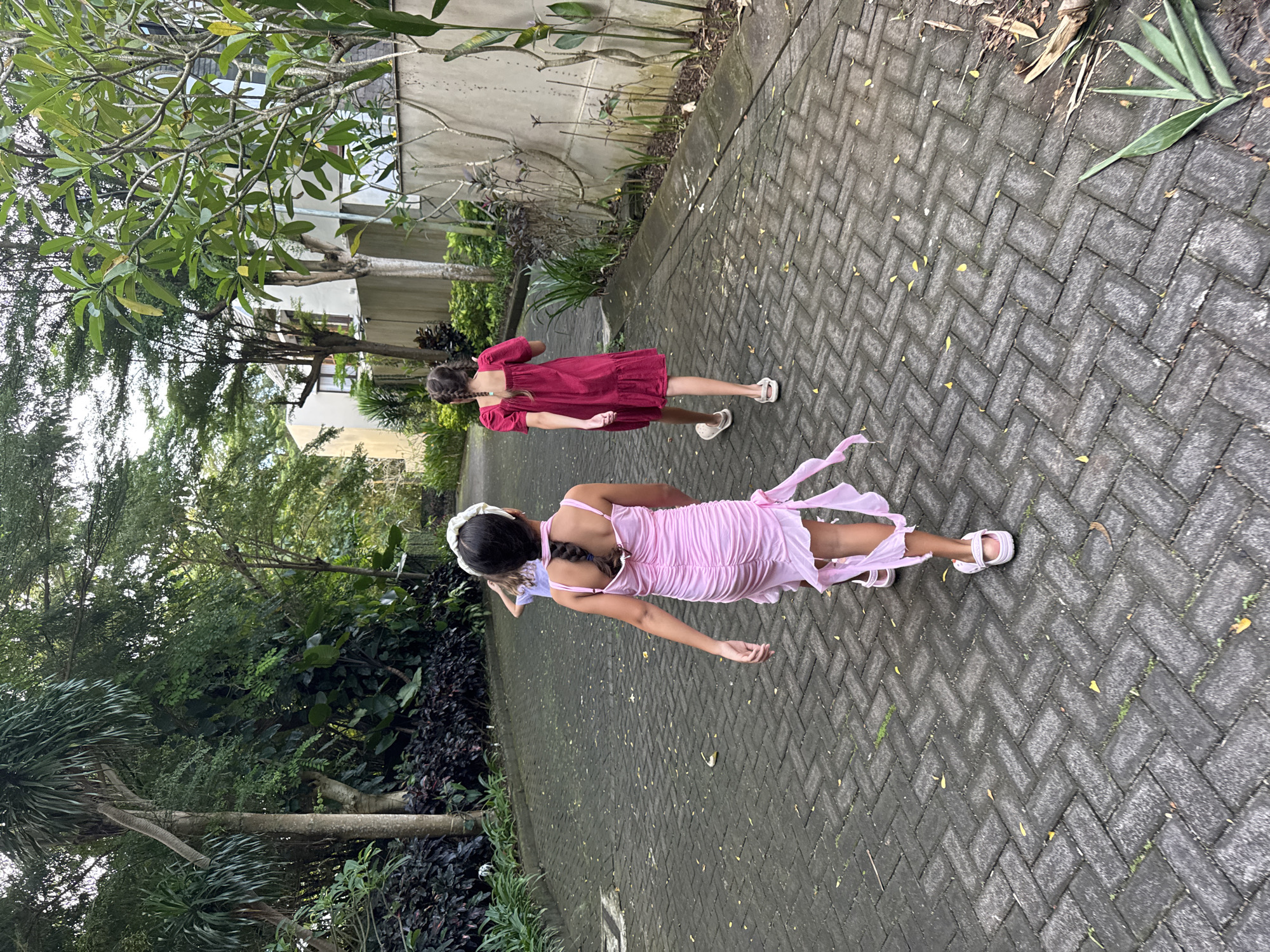 Two girls are depicted walking away from the camera on a paved pathway in Kuta Selatan, Indonesia. The path is constructed from grey interlocking pavers laid in a herringbone pattern, with scattered yellow leaves and small patches of grass visible.
In the foreground, on the left side of the path, a girl wears a light pink, ruched, spaghetti-strap dress with sheer fabric details or ties on the back. She has dark braided hair and a white headband, and is wearing white open-toed sandals. Further along the path, on the right, a second girl is seen wearing a short-sleeved, maroon or dark red tiered dress. She also has dark braided hair and wears white closed-toe sandals or clogs.
The pathway is surrounded by lush, tropical foliage, including large trees with dense green canopies, slender palm trees, and various bushes and plants. Partial views of light-colored buildings are visible through the trees in the background, suggesting a developed area within a natural setting. The scene is brightly lit, indicating daytime, possibly under overcast conditions given the diffused light and lack of harsh shadows. No visible text is present in the image.