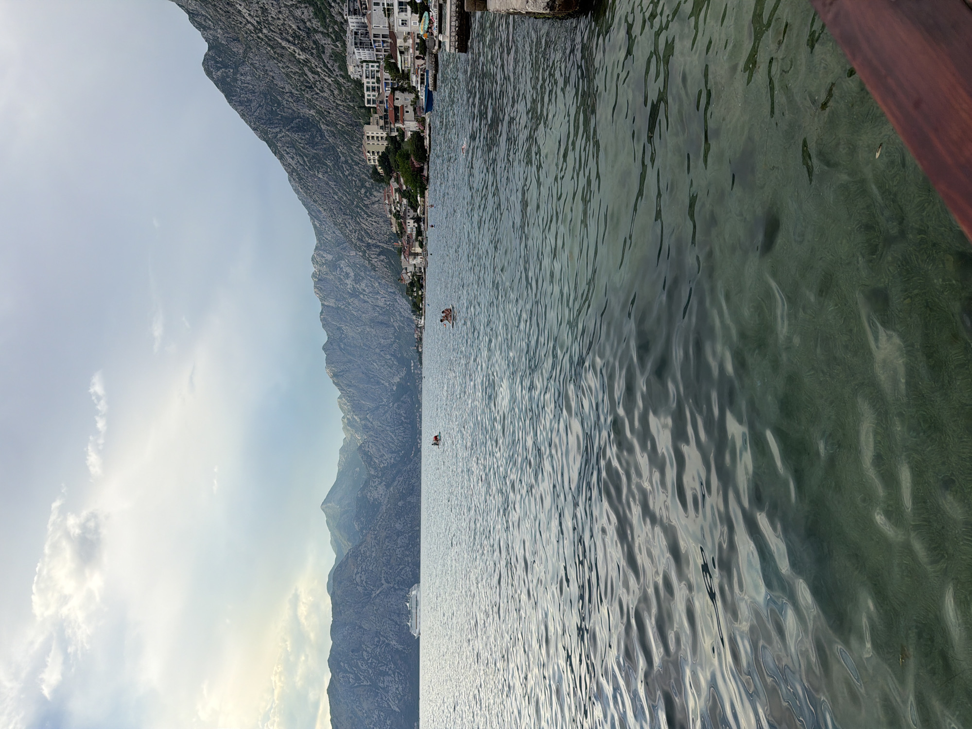 The image shows a body of water with several people swimming and relaxing. In the background, there are mountains with a town built on their slopes. The town consists of white and red-roofed buildings. A large cruise ship is visible on the water in the distance. The water is clear enough to see the bottom in some areas. The sky is partly cloudy. The overall scene suggests a coastal or bay area, likely a tourist destination given the presence of a cruise ship and swimmers. This view aligns with the typical scenery found in Dobrota, Montenegro, known for its picturesque waterfront and proximity to steep, mountainous terrain.